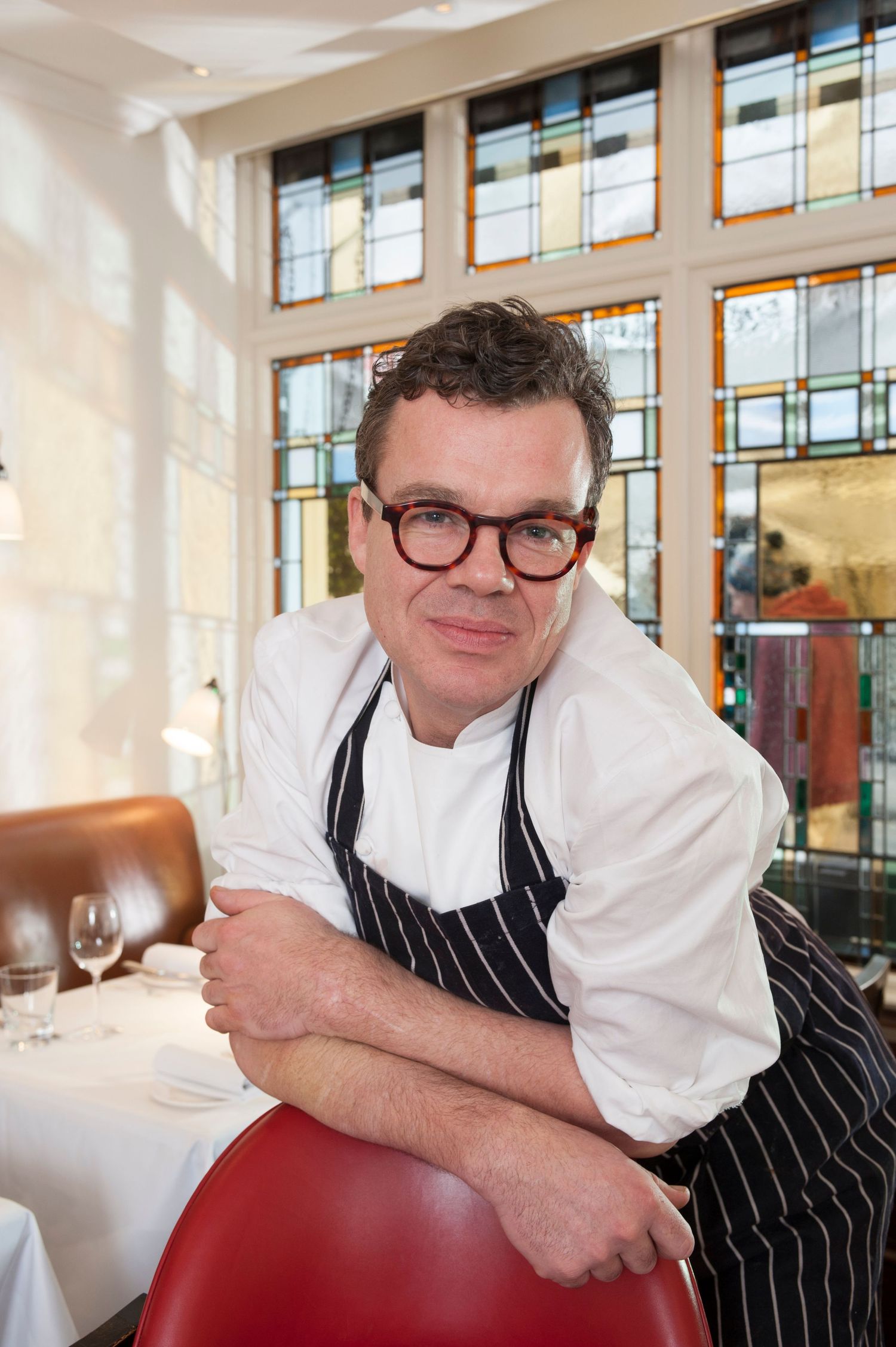 Chef in white jacket and striped apron sits smiling in elegant restaurant with stained glass windows.