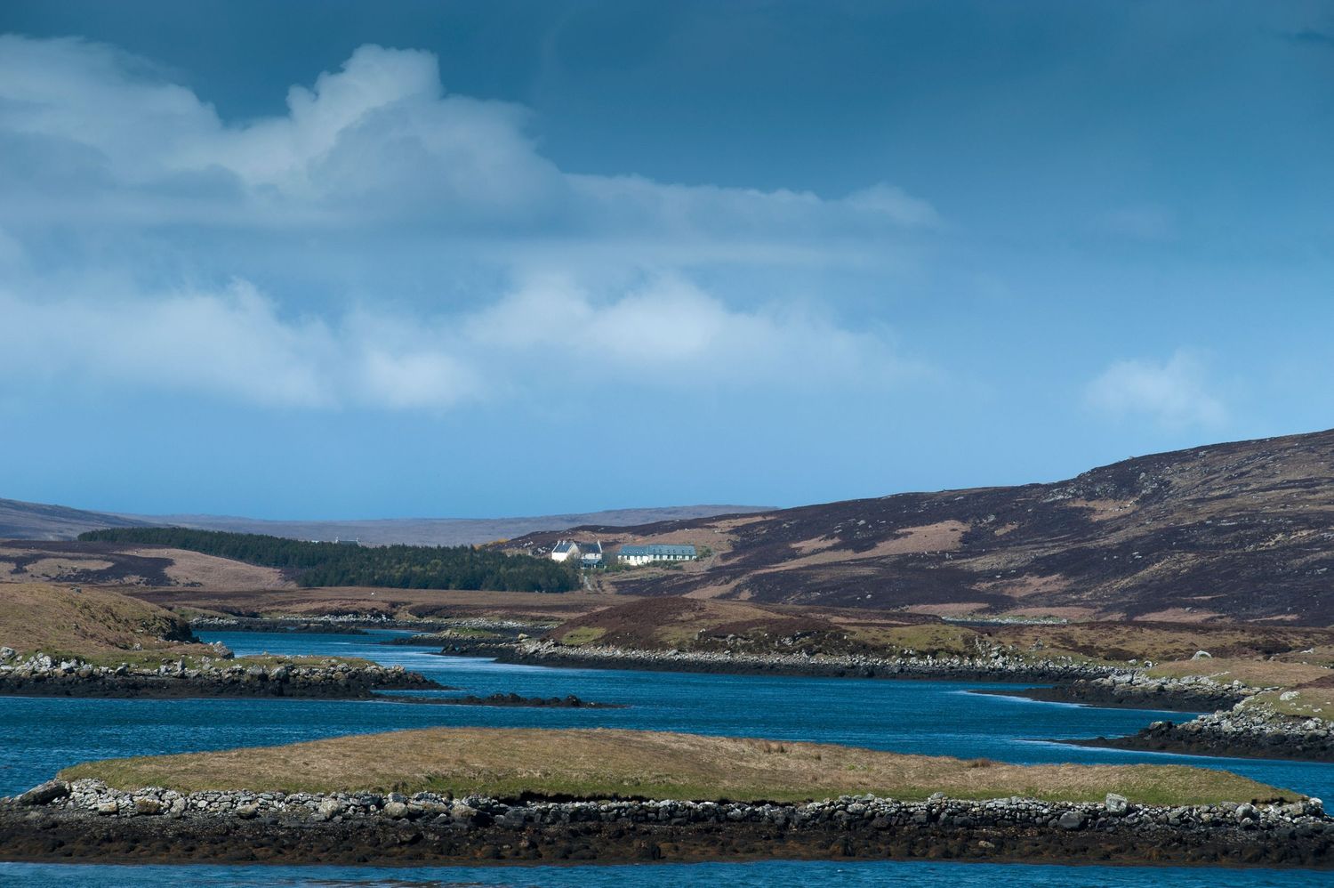 Scenic Scottish loch landscape with blue waters, rolling hills, and a white building under dramatic cloudy skies.
