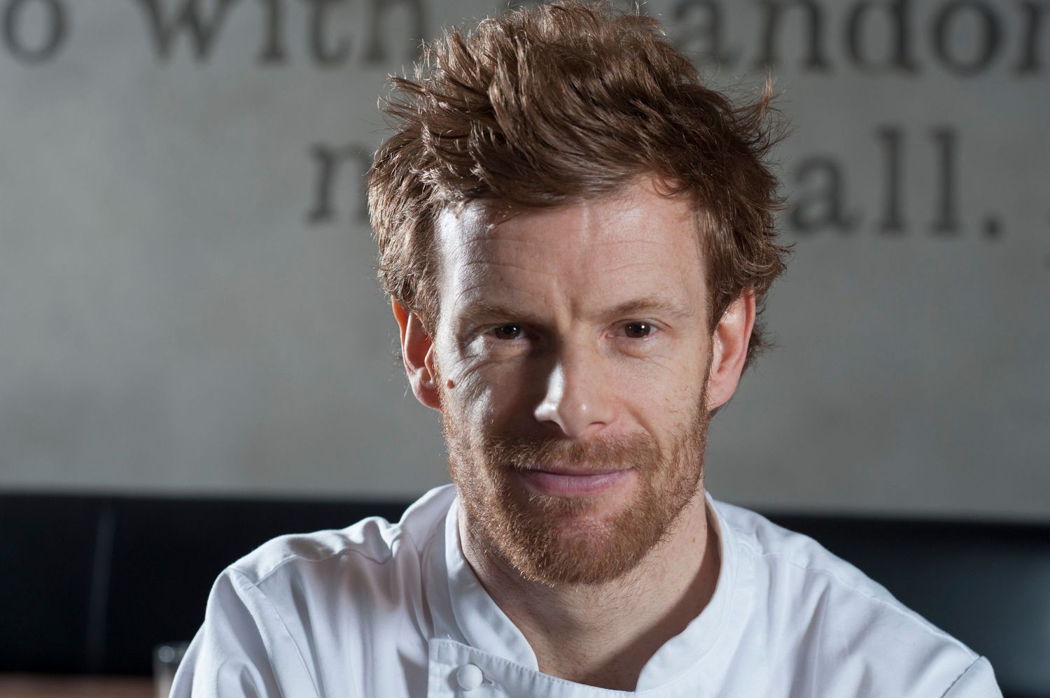 A red-haired male chef in a white uniform smiles confidently in a modern restaurant setting.