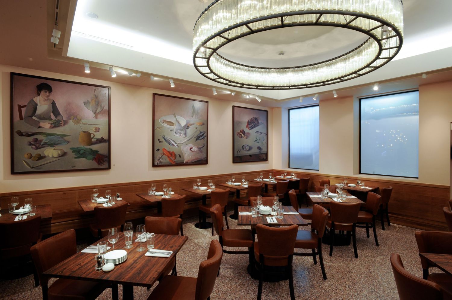 Elegant restaurant dining room with round chandelier, brown leather chairs, and large artwork panels on wood-paneled walls.