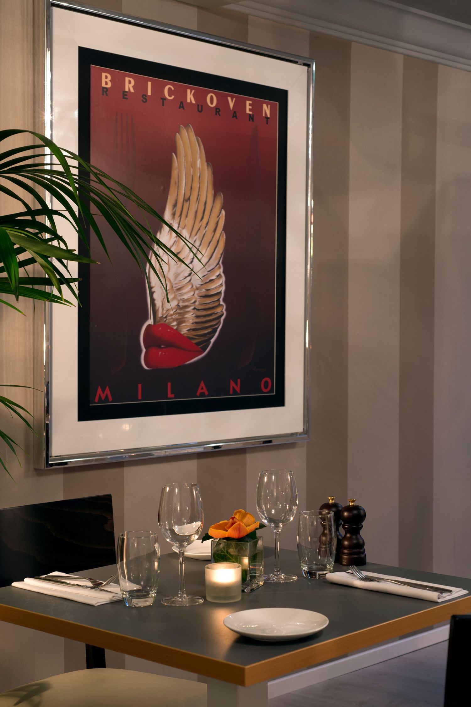Brickoven Milano restaurant interior with elegant table setting, wine glasses, candle, and framed winged logo poster on wall.