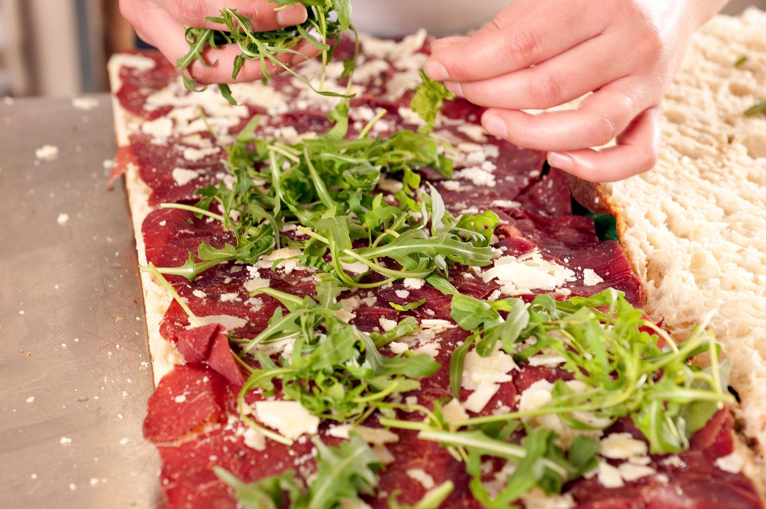 Hands sprinkling fresh arugula over a flatbread topped with red sauce, ricotta, and herbs on a floured surface.