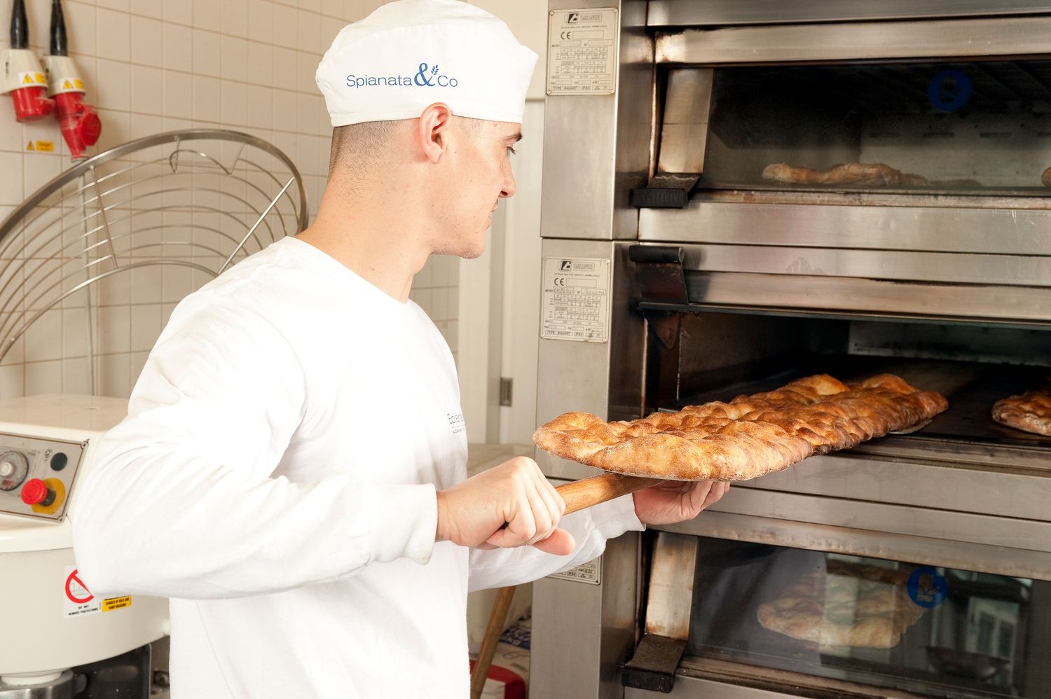 Baker in white uniform removing freshly baked bread from a commercial deck oven in a professional bakery.