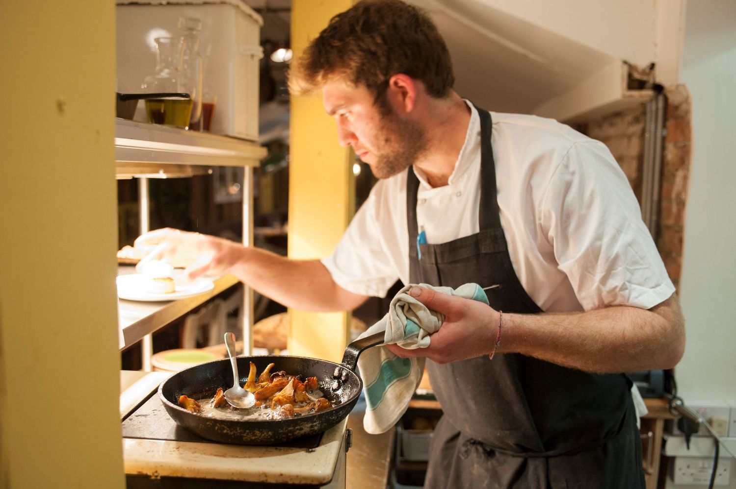 A bearded male chef in apron cooking in a skillet on a stovetop in a professional kitchen.