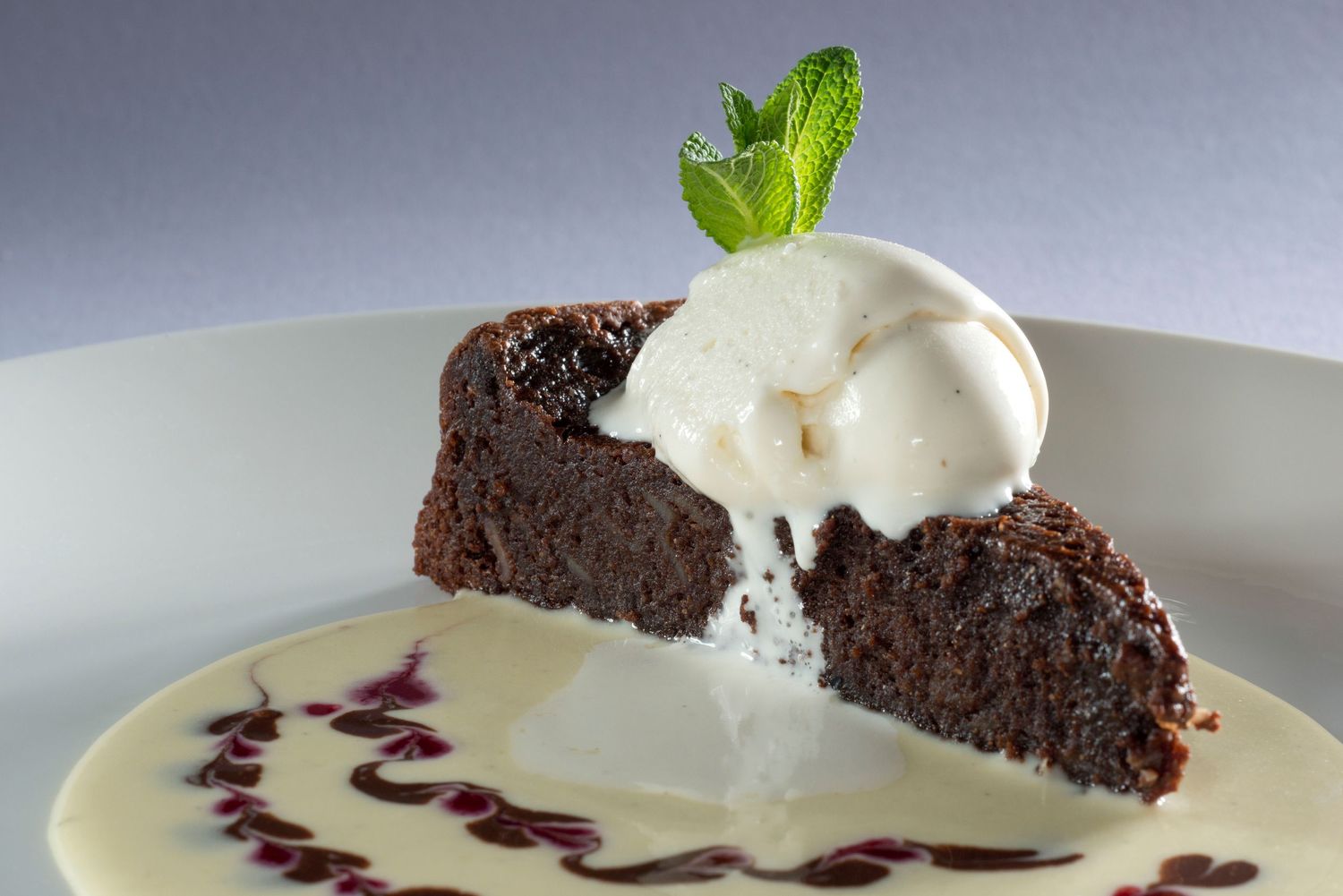 Rich chocolate brownie slice topped with vanilla ice cream and mint, served on a white plate with berry sauce.
