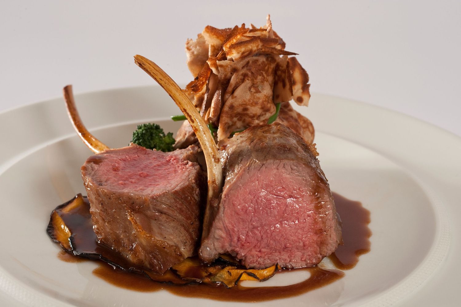 Elegant rack of lamb with pink medium-rare chops, rich brown sauce, broccoli, and crispy garnish on a white plate.