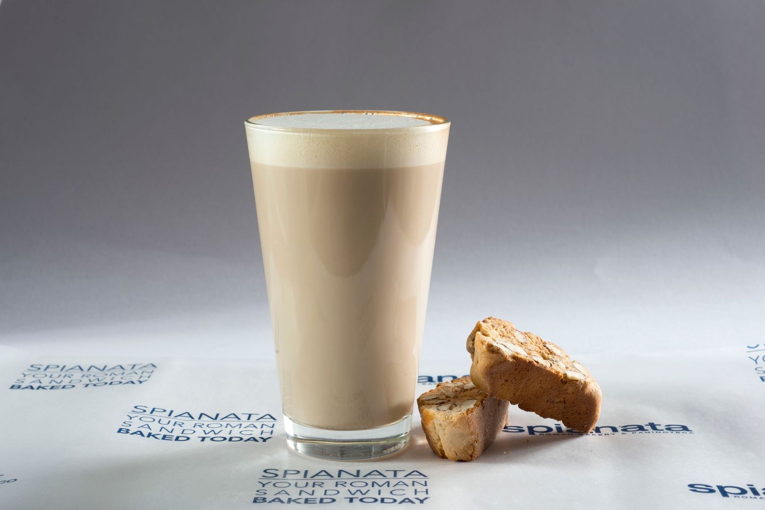 A tall glass of creamy latte beside two pieces of biscotti on a branded Spianata paper.