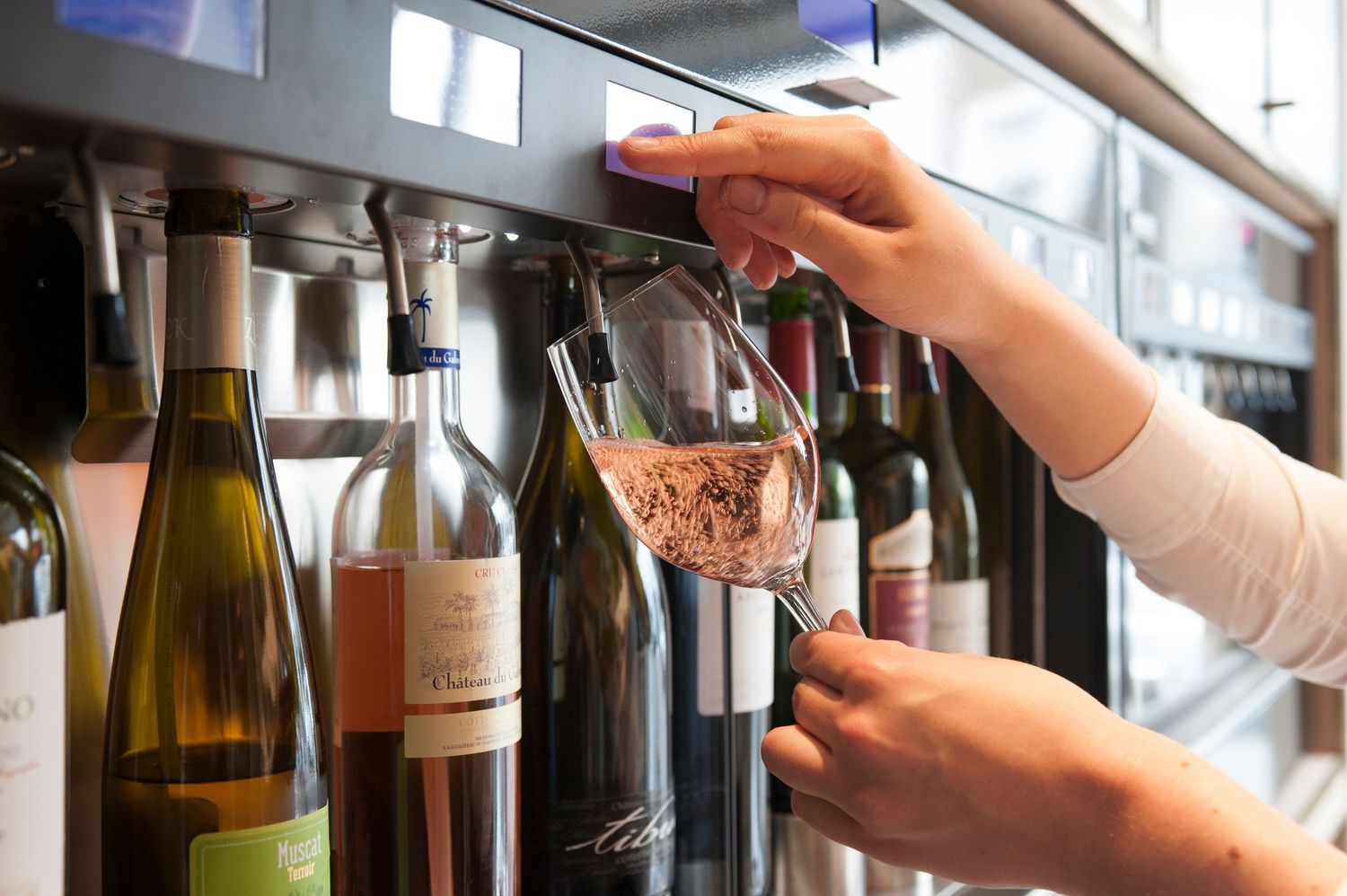 Person using a wine dispenser machine to pour rose wine into a glass surrounded by bottles.