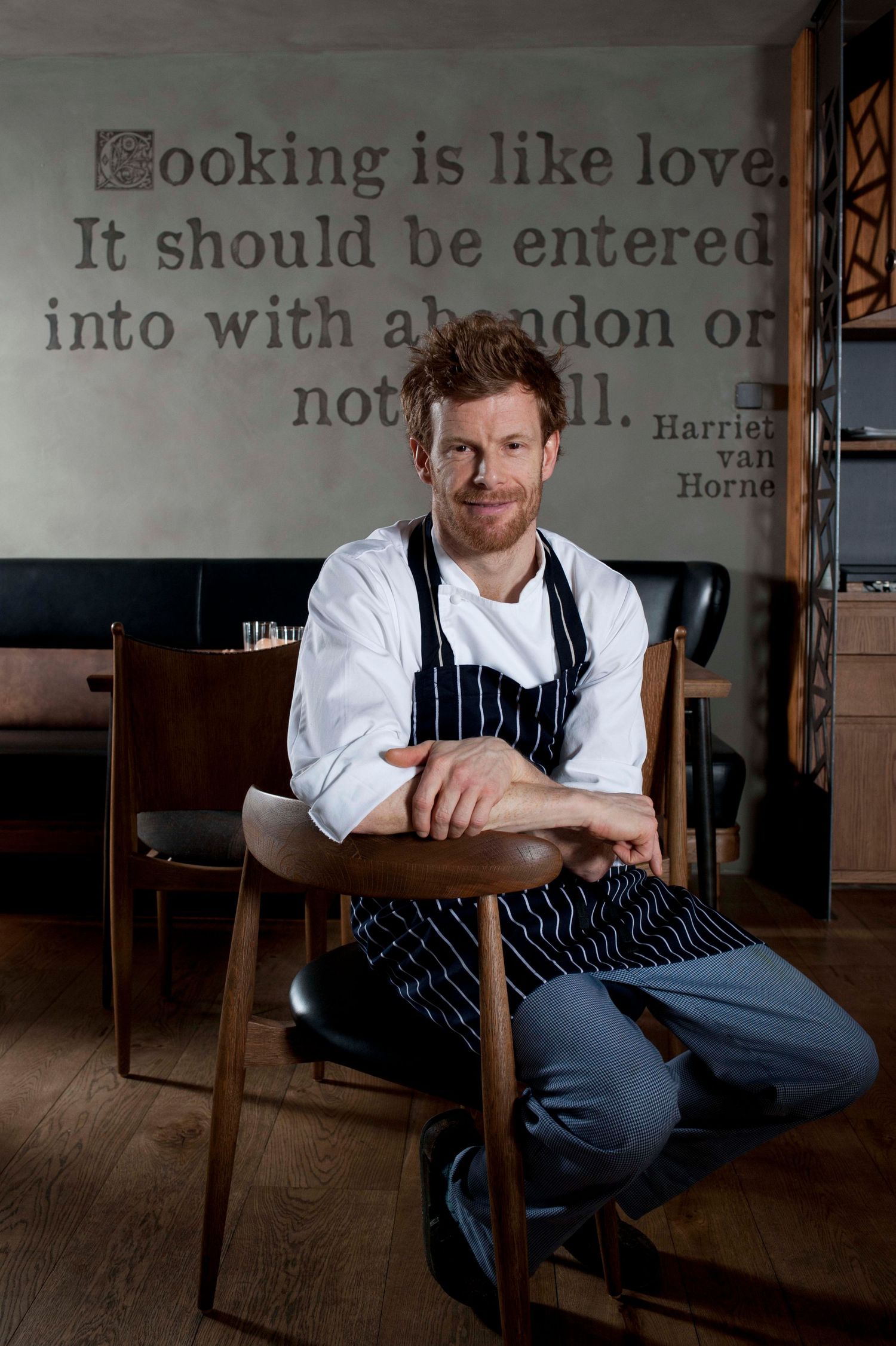 A smiling chef in white jacket and striped apron sits relaxed on a chair in a restaurant with a cooking quote on the wall.