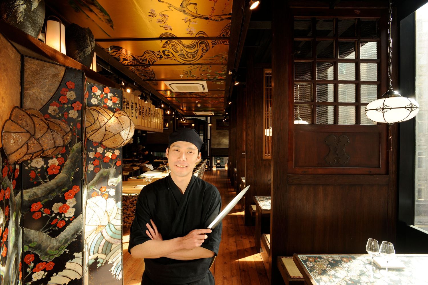 A smiling chef in black uniform holds chopsticks in a traditional Japanese restaurant with ornate wooden decor and floral wall art.