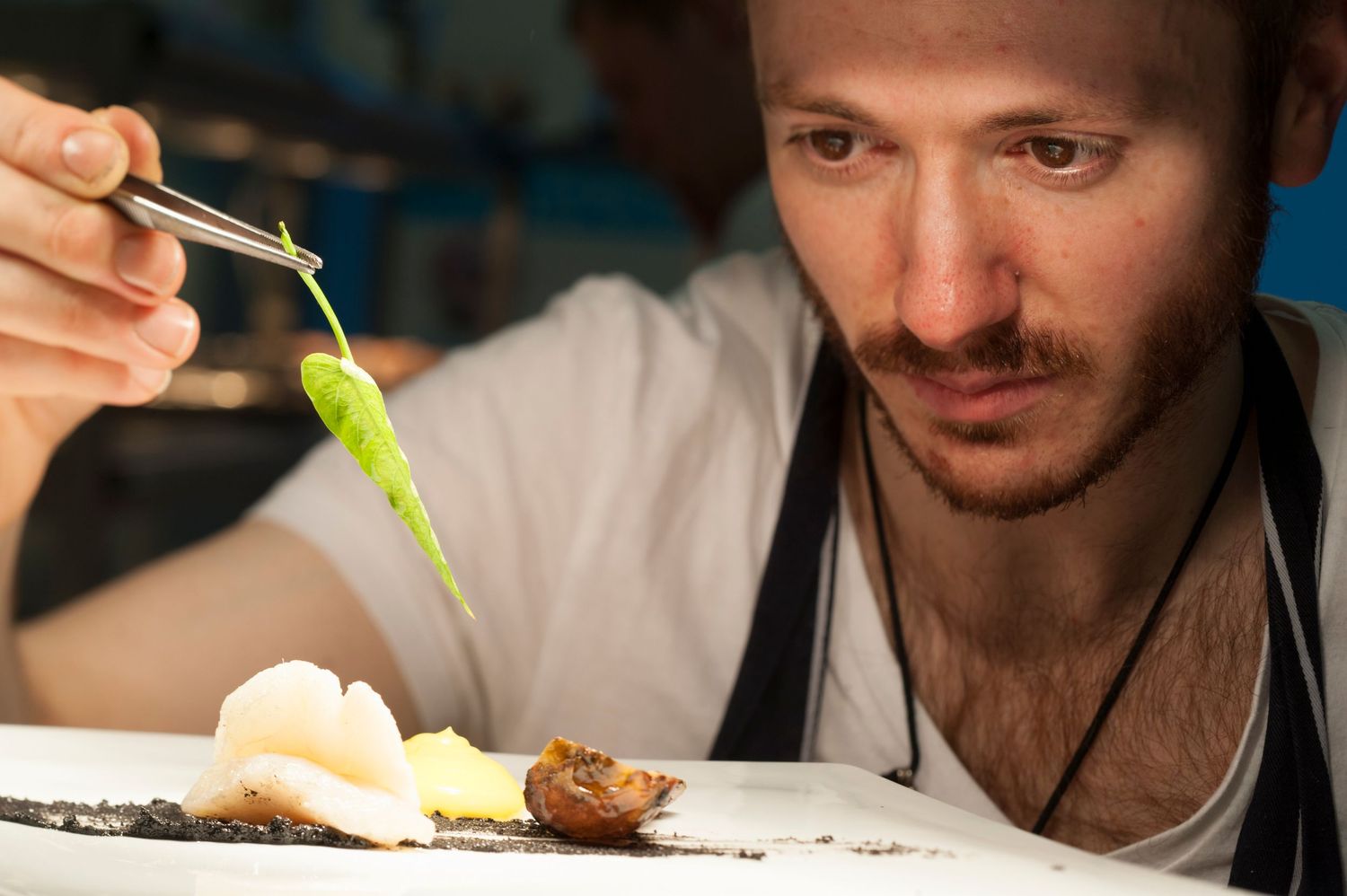 Chef carefully plates a gourmet dish using tweezers, adding a green garnish to an elegant white plate.