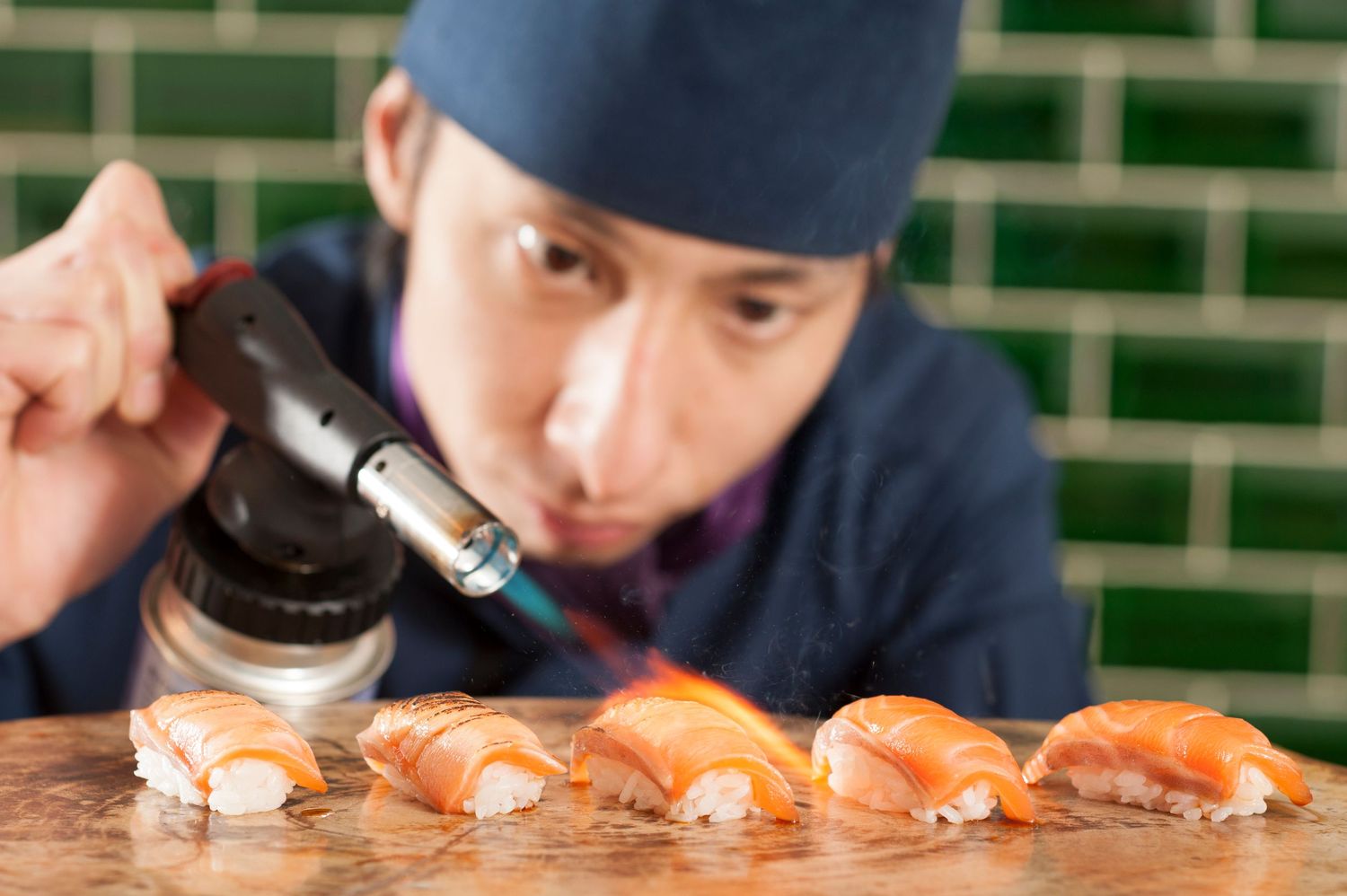 A Japanese chef uses a blowtorch to sear fresh salmon nigiri sushi on a marble counter in a restaurant kitchen.