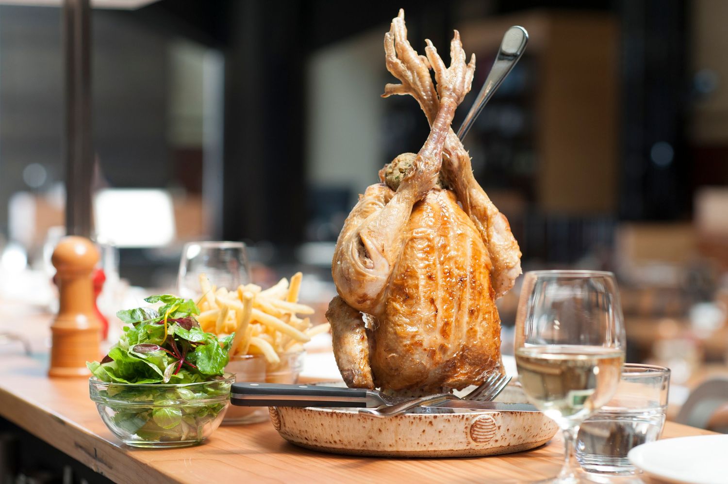 Whole roasted chicken served upright on a tray with golden french fries and fresh green salad in a restaurant setting.