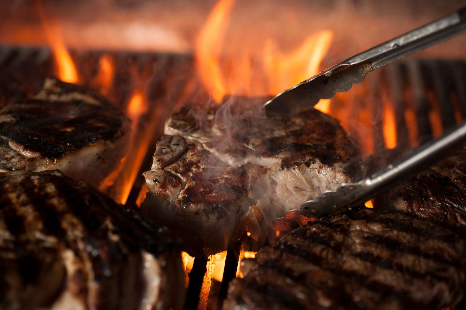Juicy steaks sizzling on a flaming grill, held with metal tongs, with smoke rising from the charred meat.