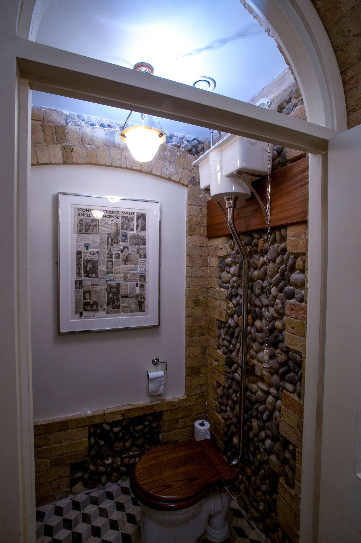 Rustic bathroom with stone walls, wooden toilet seat, high cistern, framed newspaper art, and checkered floor tiles.