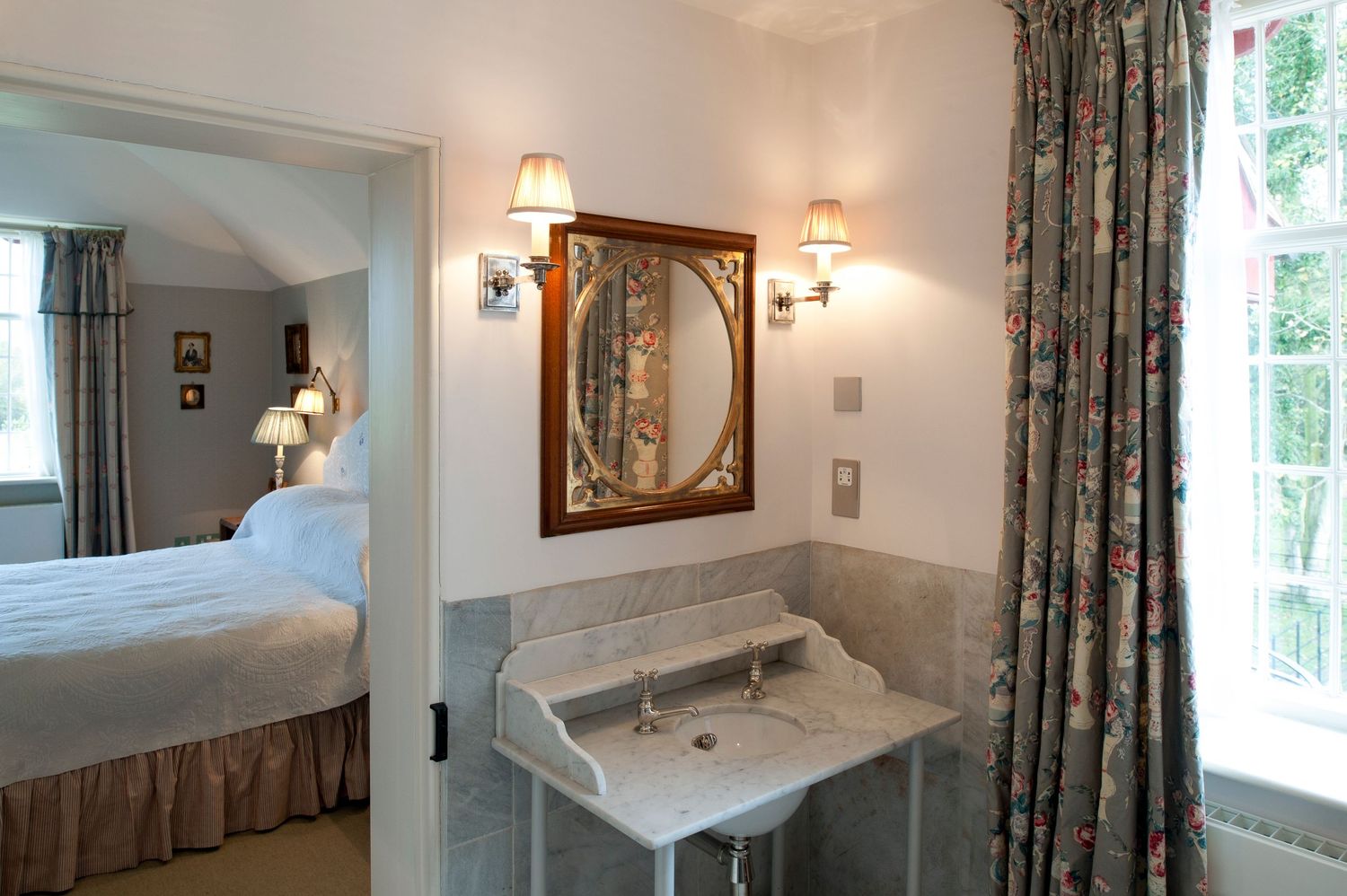 Elegant hotel room with marble pedestal sink, ornate mirror, wall sconces, floral curtains, and a glimpse of a cozy bedroom.
