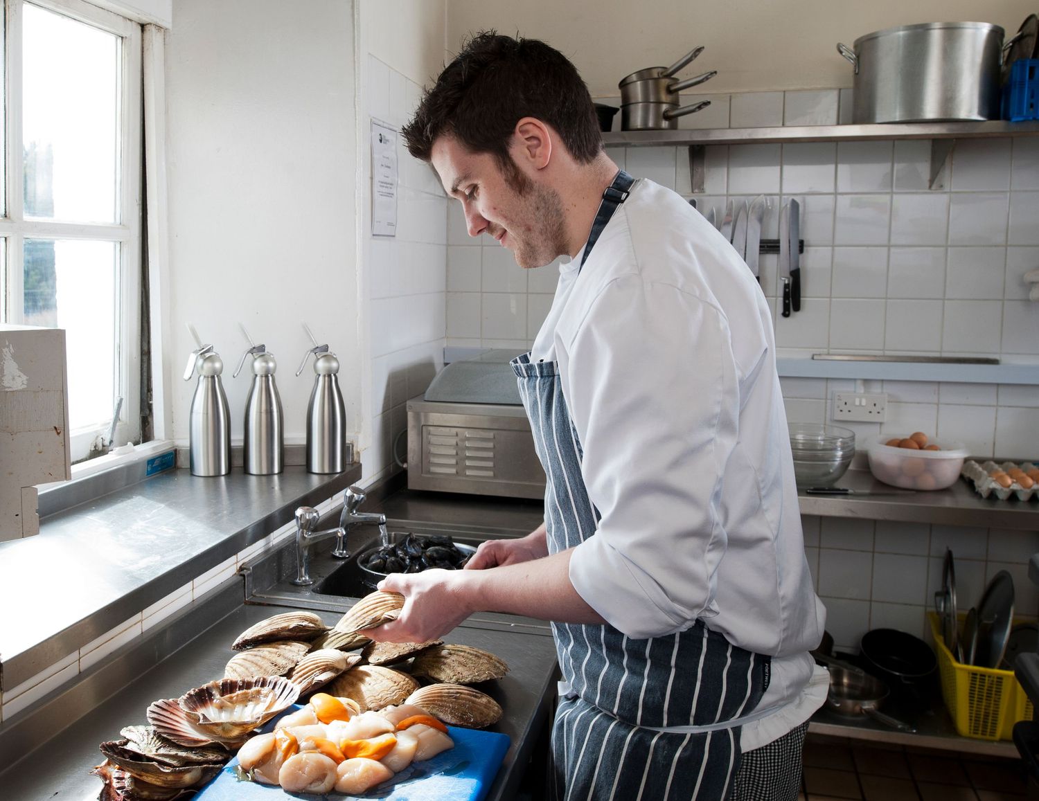Chef in striped apron arranging fresh bread and pastries on a kitchen counter in a professional restaurant kitchen.