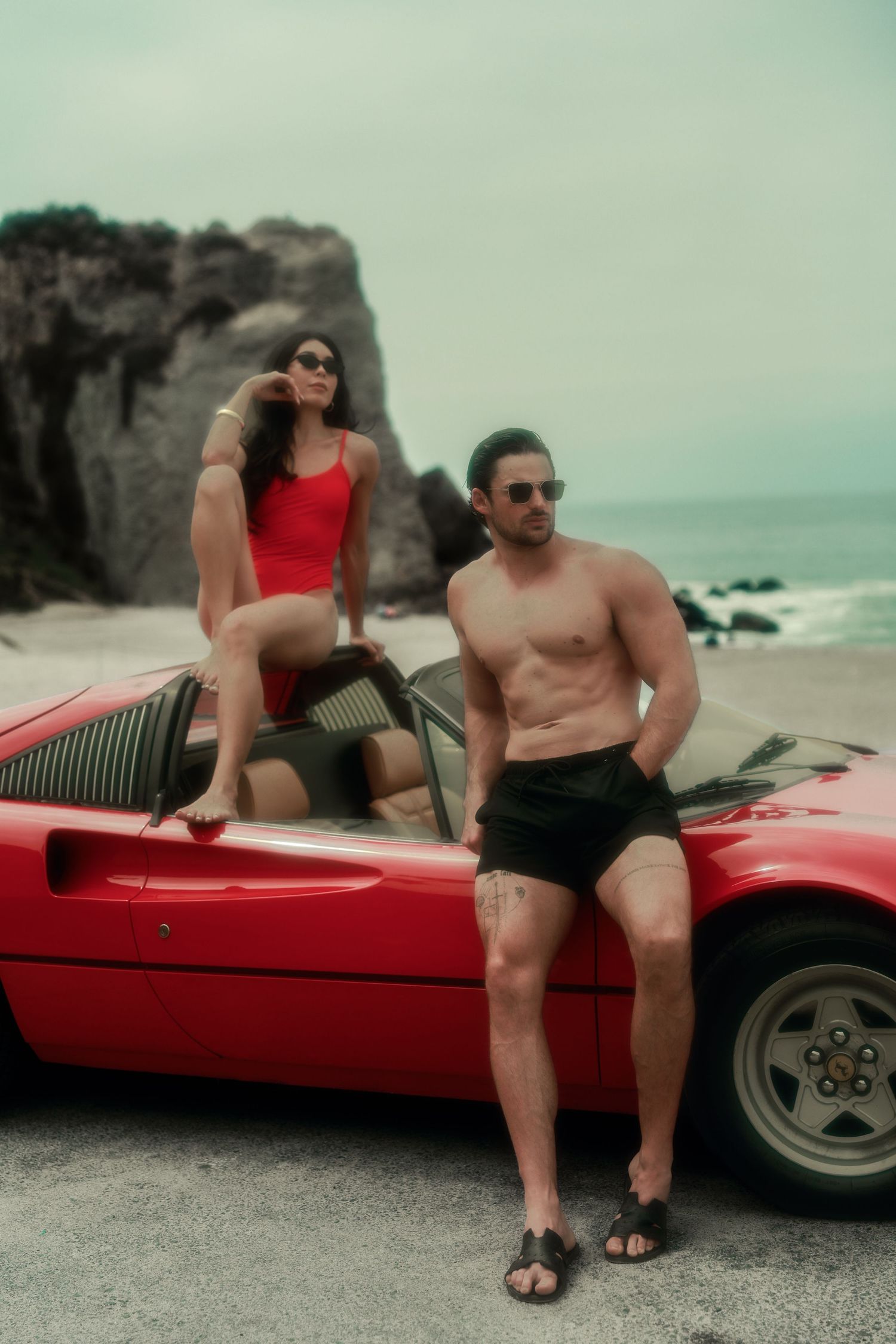 A couple poses with a red Ferrari convertible on a rocky beach, woman in red swimsuit, man shirtless in black shorts.