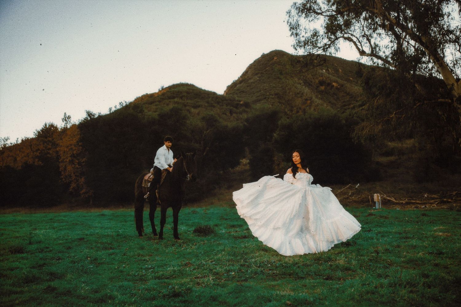 Bride in flowing white gown twirls in green field as groom rides black horse with lush hills behind.
