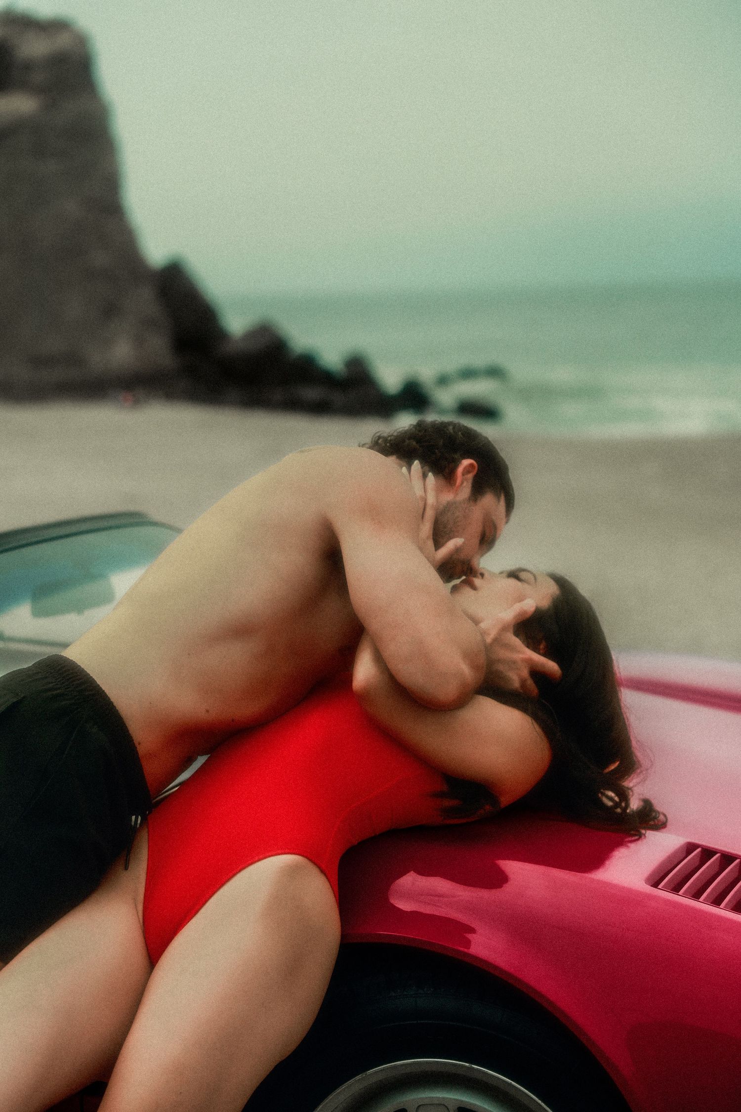 Romantic couple kissing on a red sports car at a misty beach, woman in red swimsuit.