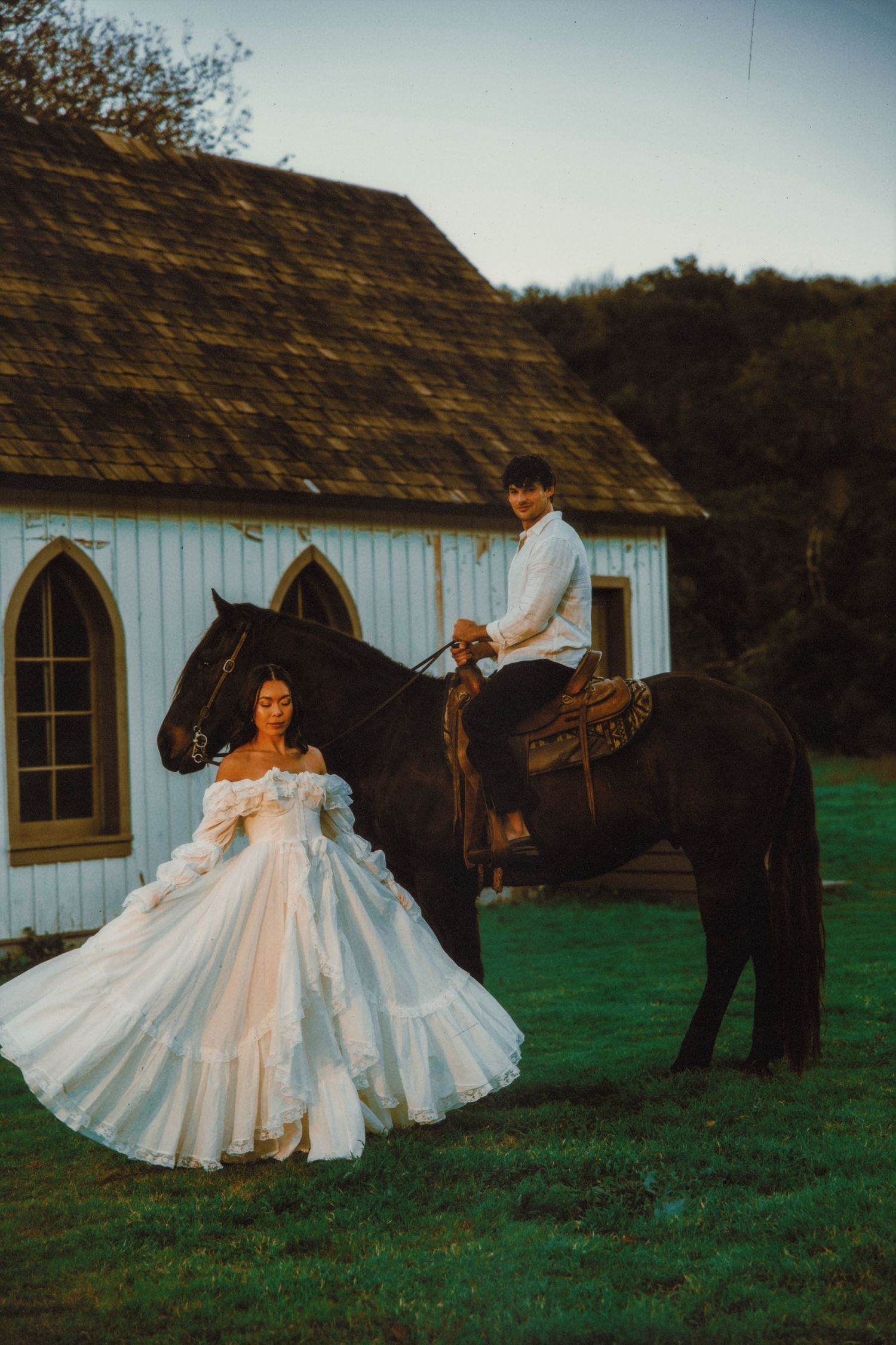 A bride in a white ball gown stands beside a dark horse ridden by a groom near a white chapel at dusk.