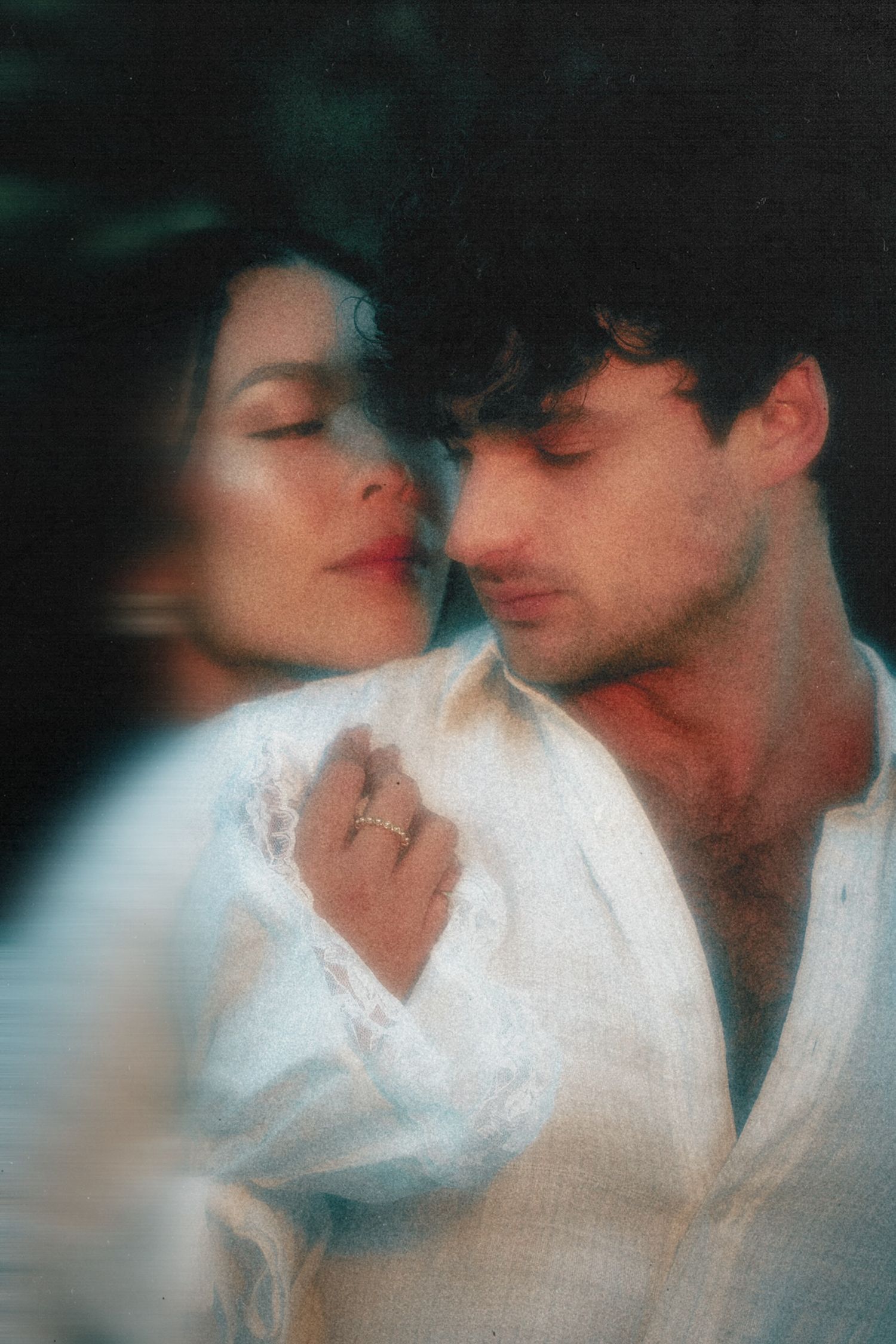 A romantic couple in soft focus, woman in white lace resting her head on a mans shoulder, both with eyes closed.