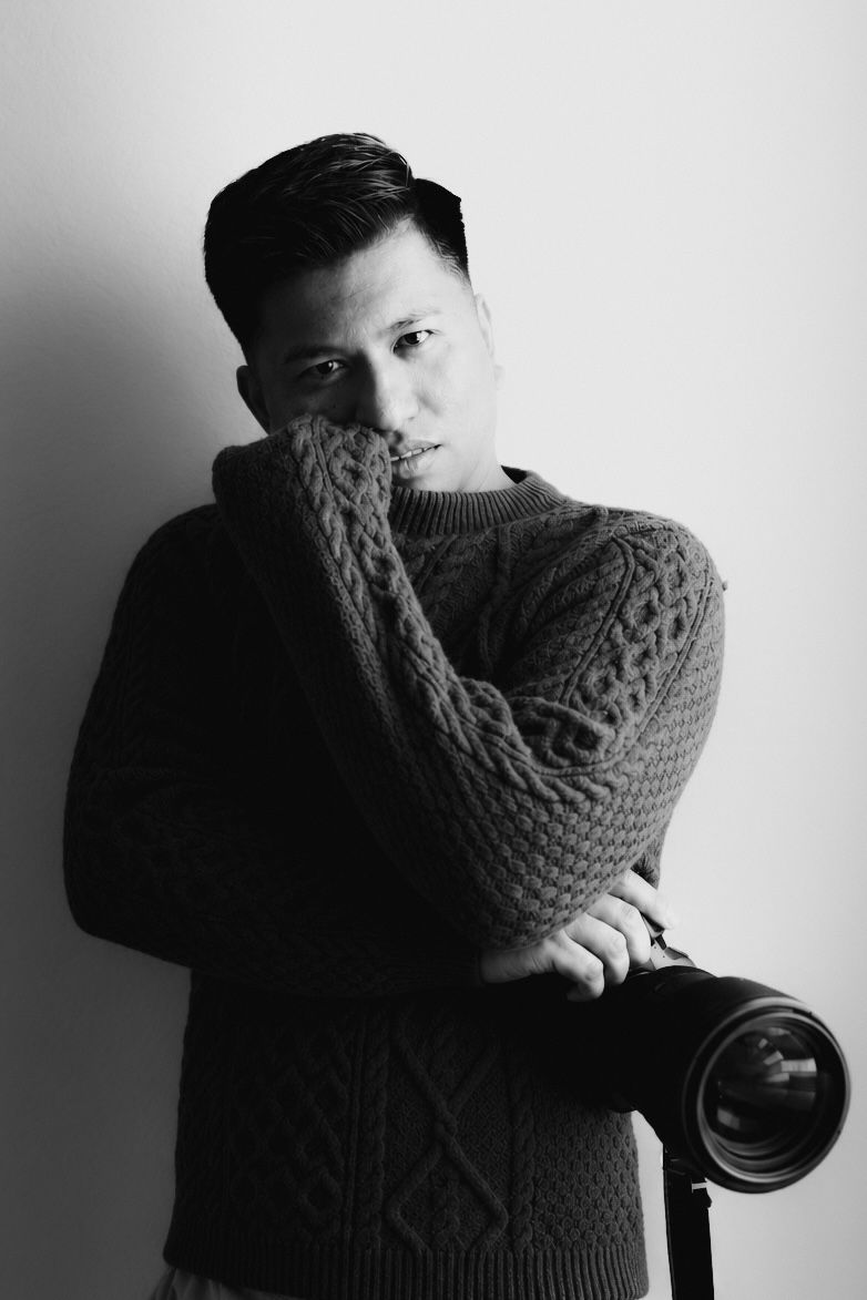 Black and white portrait of a young Asian man in a cable-knit sweater holding a professional camera.