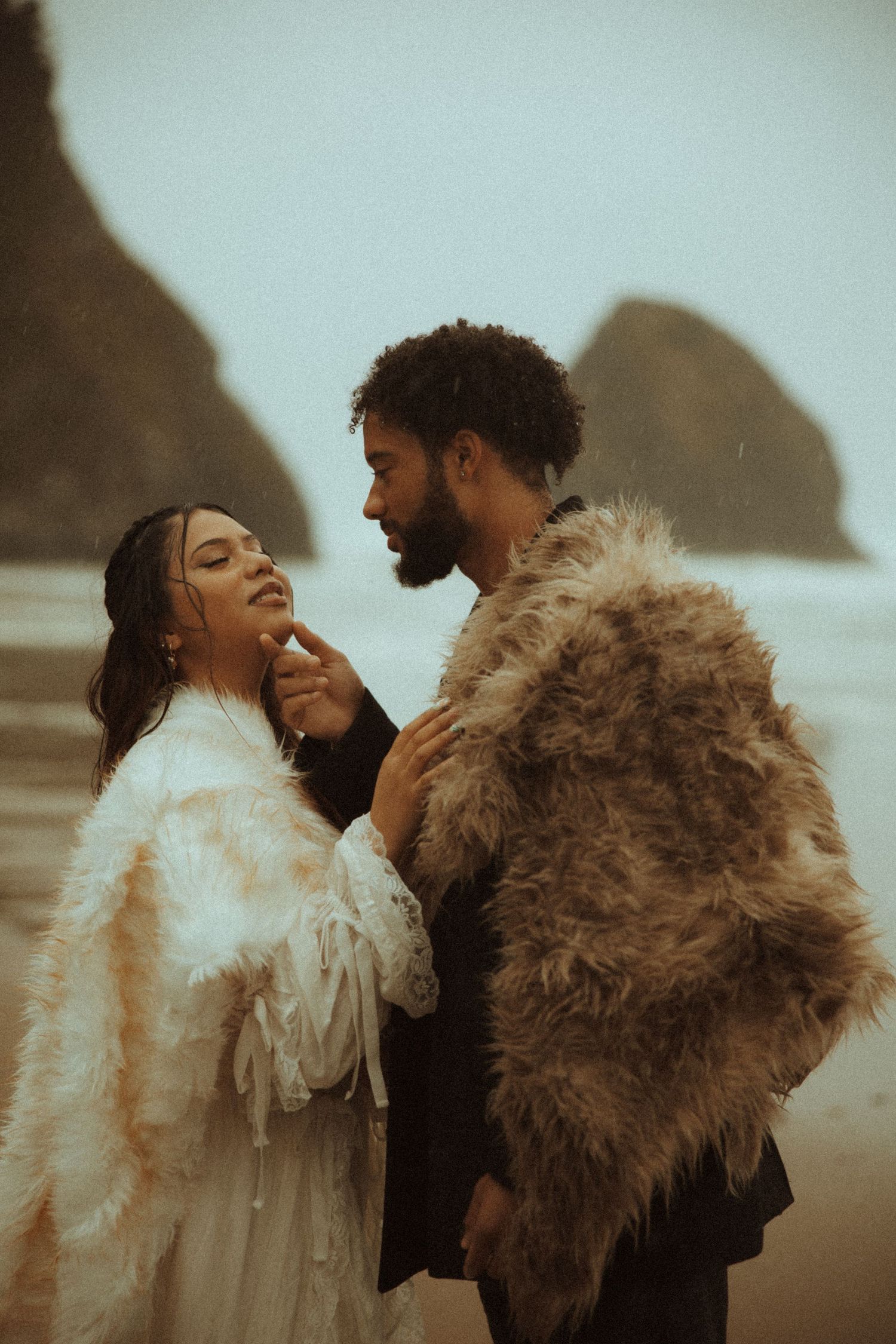 A couple in fur wraps share a romantic moment on a misty beach with dramatic sea stacks in the background.