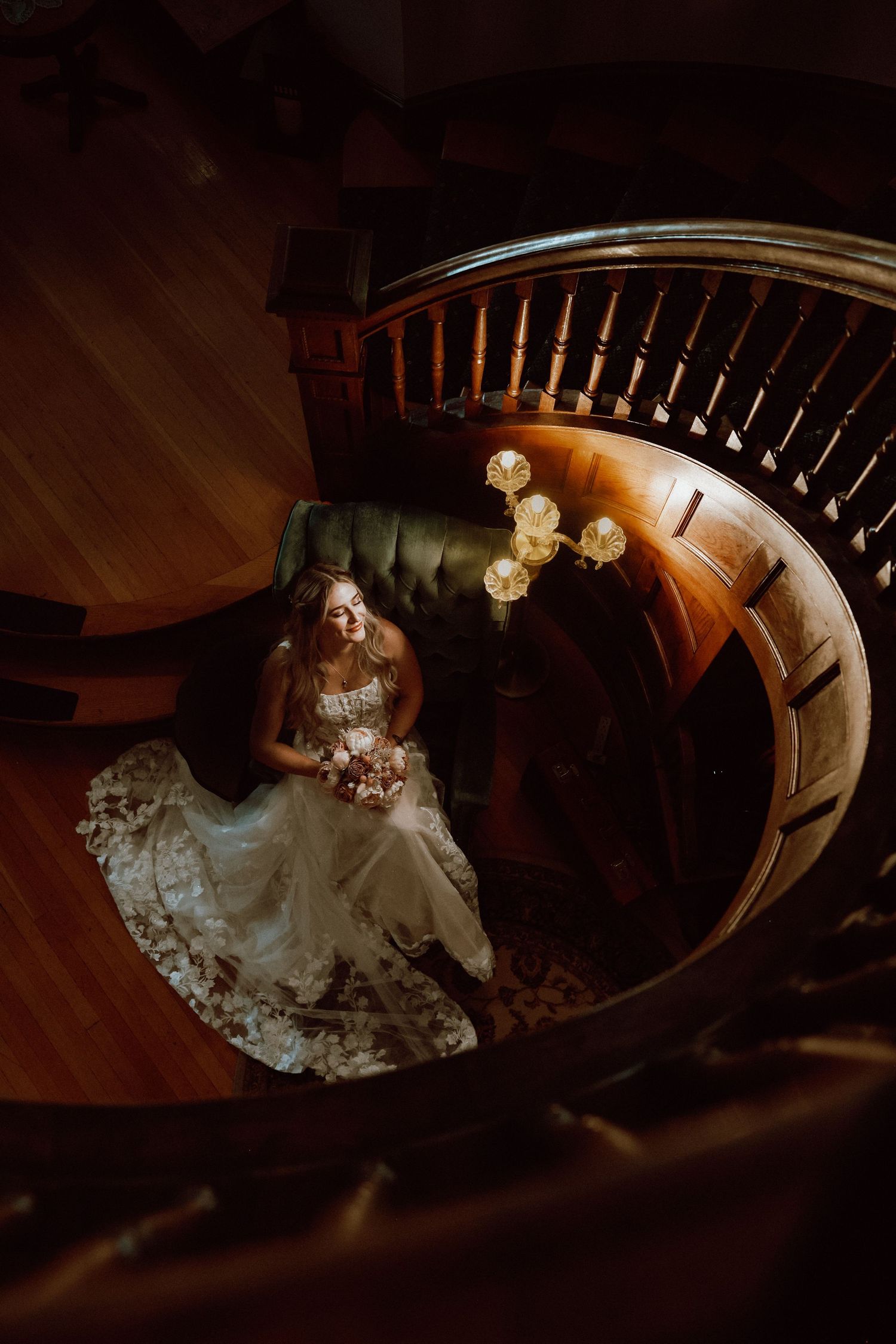 Bride in lace wedding gown holding bouquet seated at base of elegant wooden spiral staircase, viewed from above.