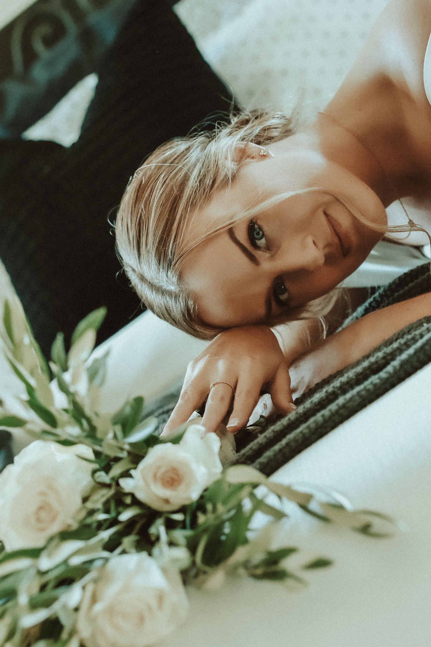 Blonde woman resting her head on a bed surrounded by white roses and green leaves, wearing a ring.