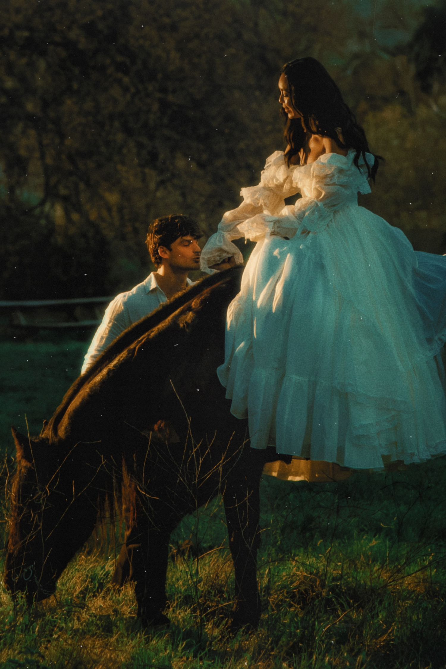 A man in white shirt kneels beside a horse, gazing up at a woman in a flowing white ball gown outdoors.