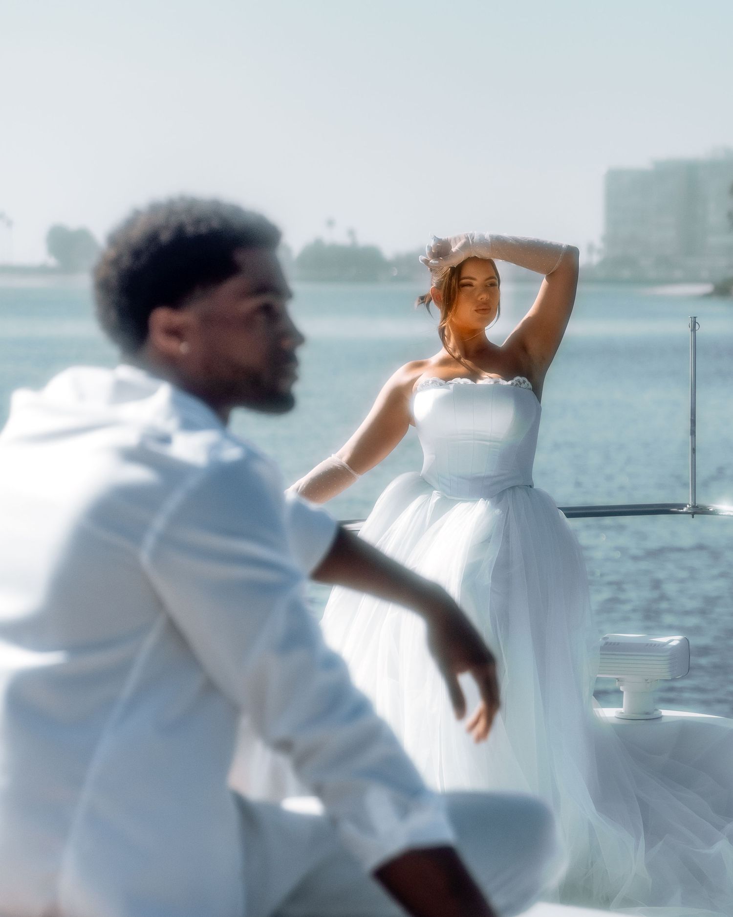 A couple in white outfits poses on a yacht with a waterfront skyline, the woman in a strapless gown looking elegant.