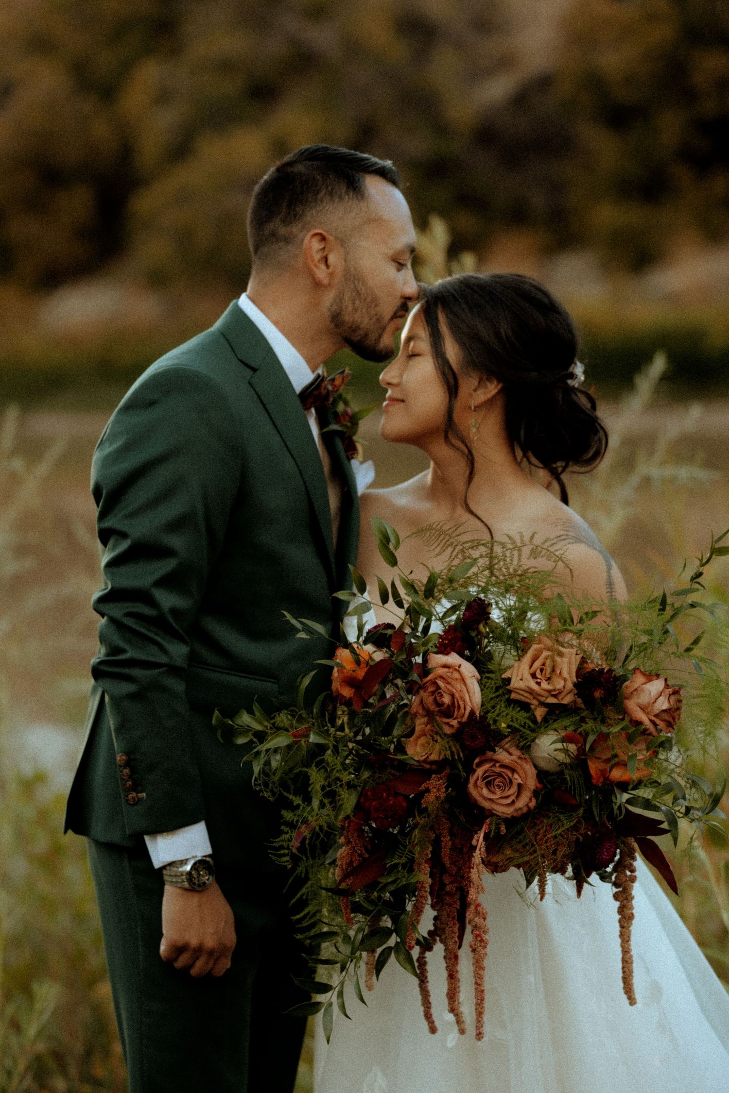 A bride and groom share a romantic moment outdoors, she holds a lush bouquet of autumn roses and greenery.