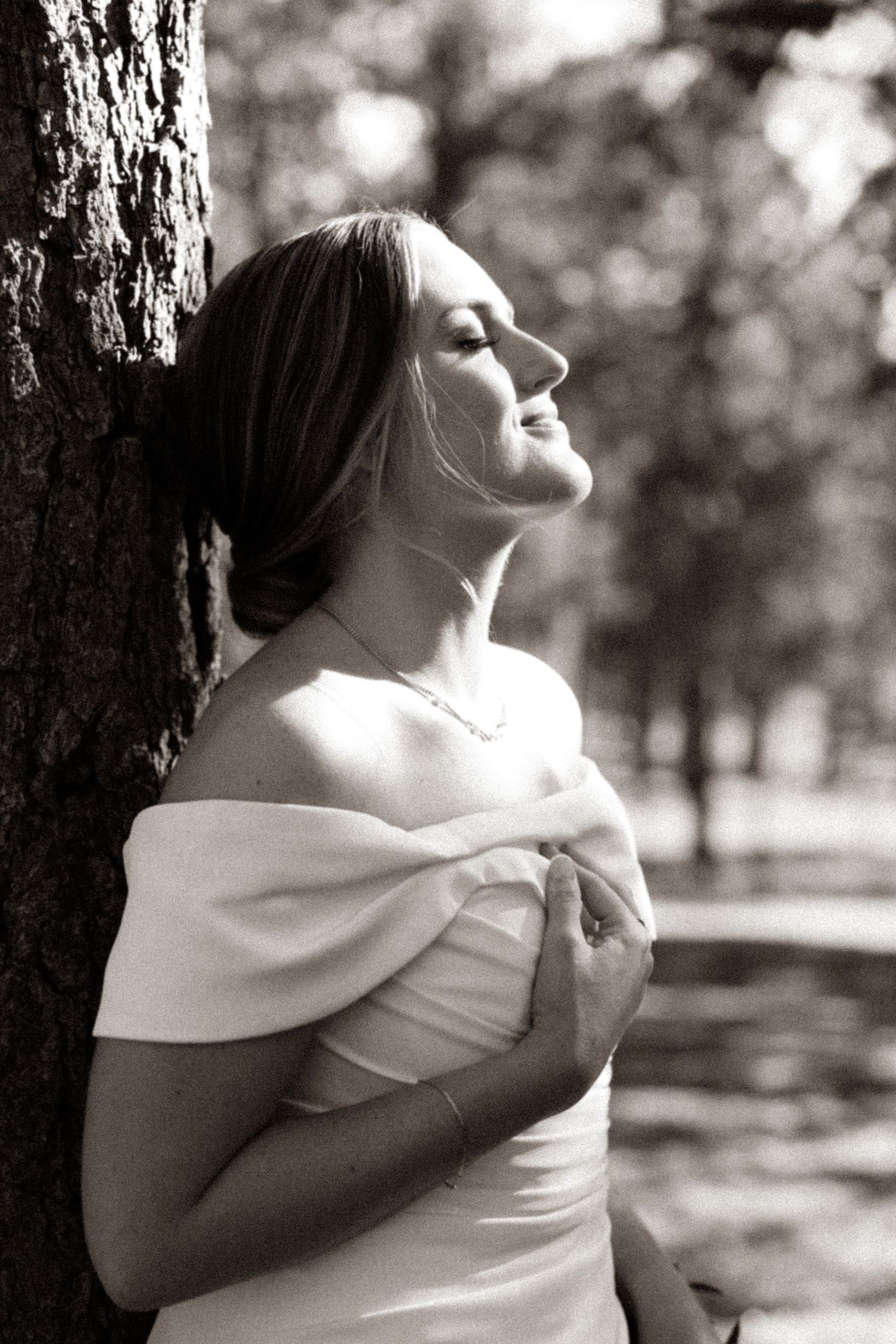 Black and white photo of a bride in off-shoulder gown leaning against a tree in a park, eyes closed, smiling.