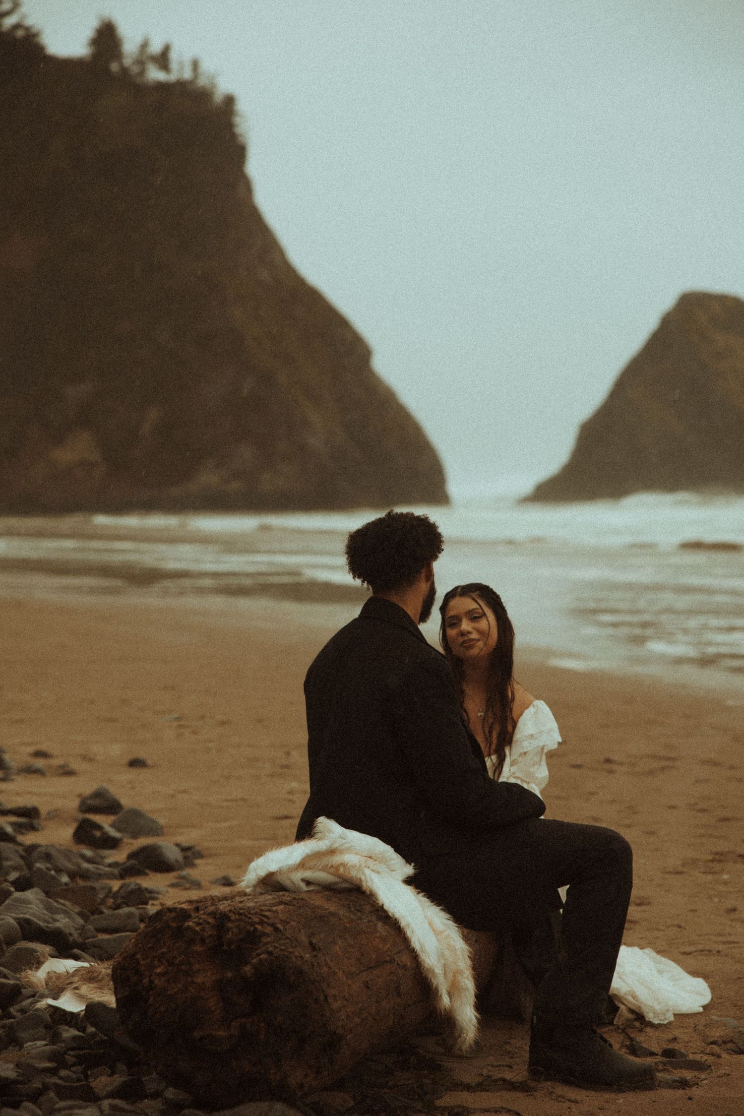 Couple sitting on driftwood log on moody Oregon coast beach with dramatic sea stacks in background.