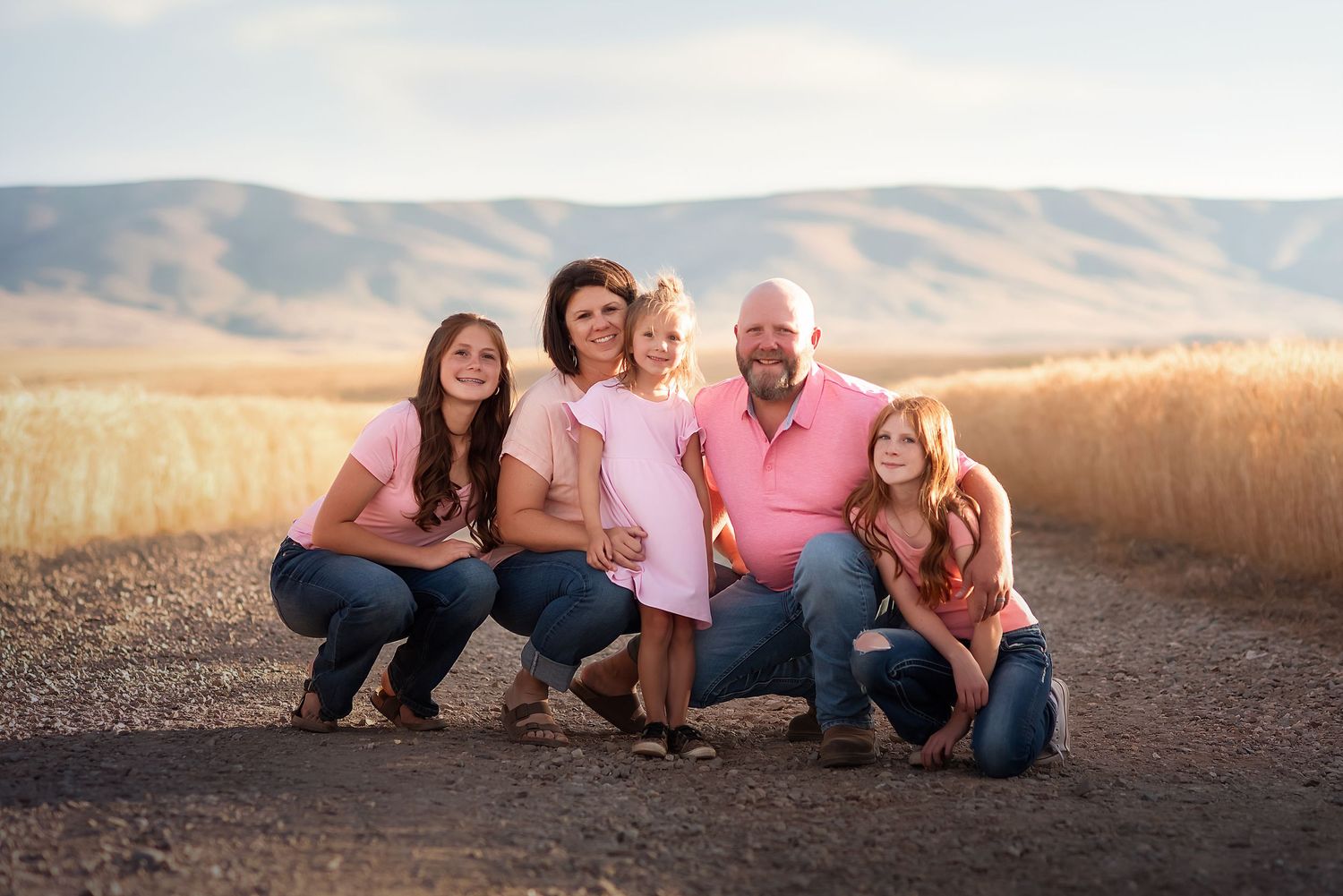 Smiling family dressed in blue jeans and pink shirts on a dirt road surrounded by golden wheat on the Waterville plateau.
