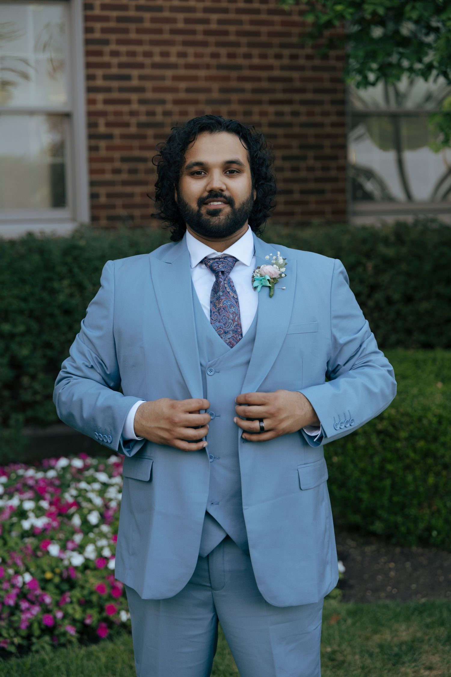 Groom in light blue three-piece suit with floral boutonniere posing confidently in garden setting.