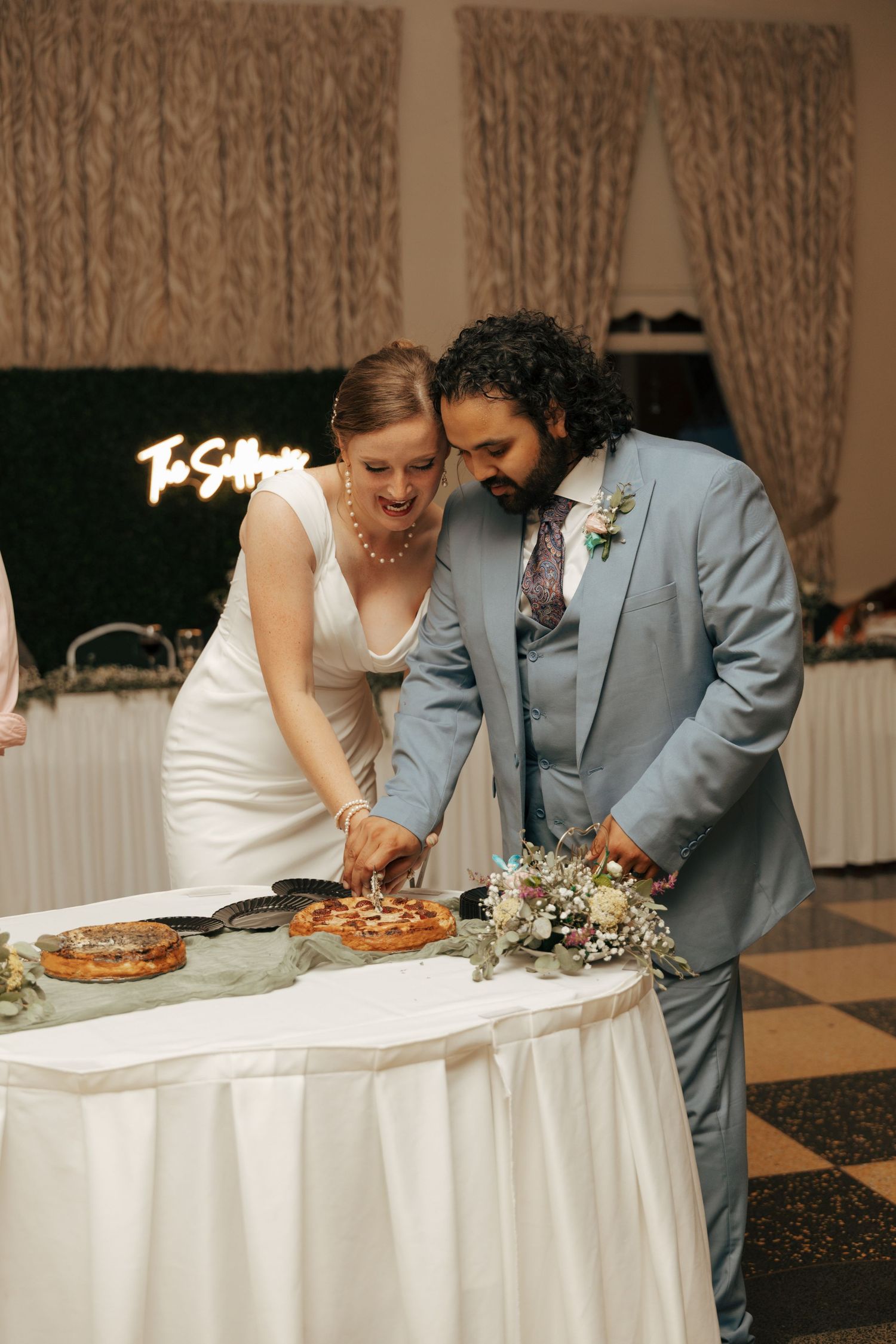 Bride and groom cutting wedding pie together at elegant reception with neon sign backdrop.