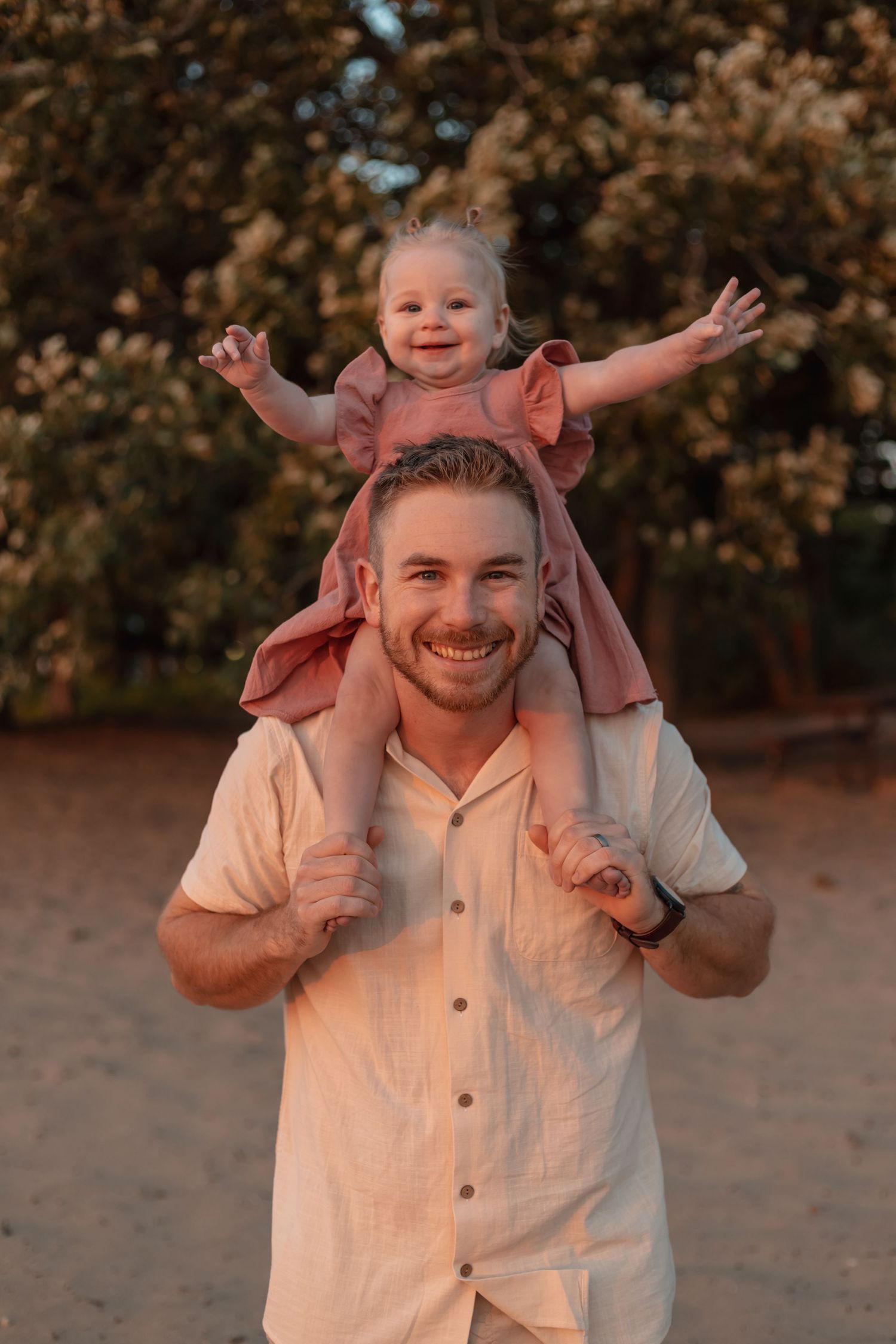 Smiling father holds toddler girl in pink dress on his shoulders outdoors at golden hour.