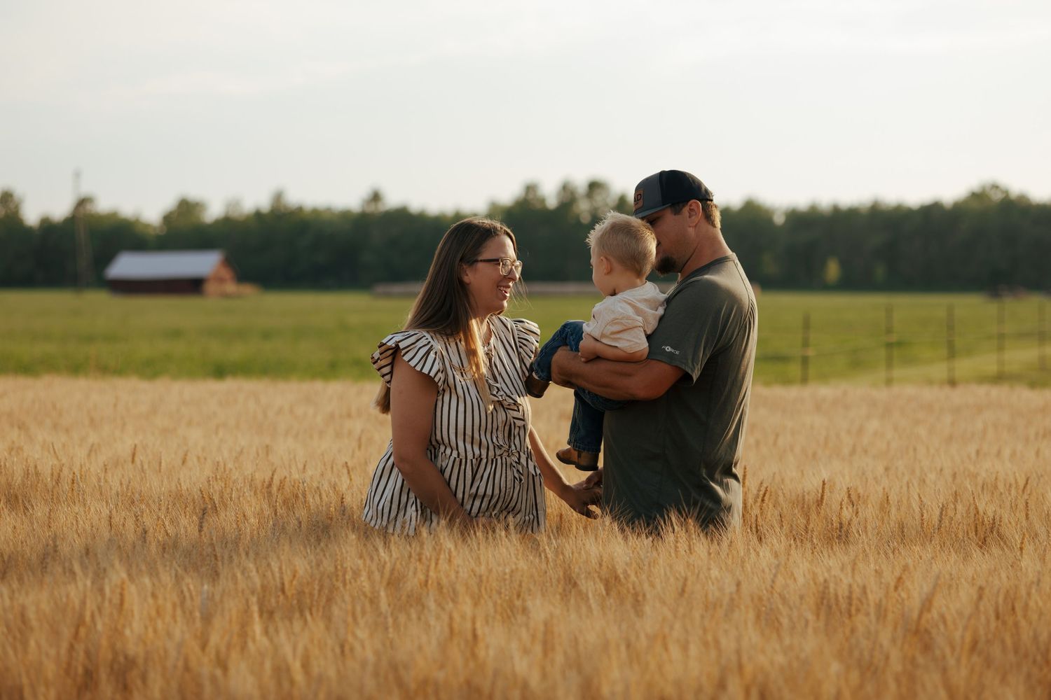 Couple with baby standing in golden wheat field at sunset near a red barn.