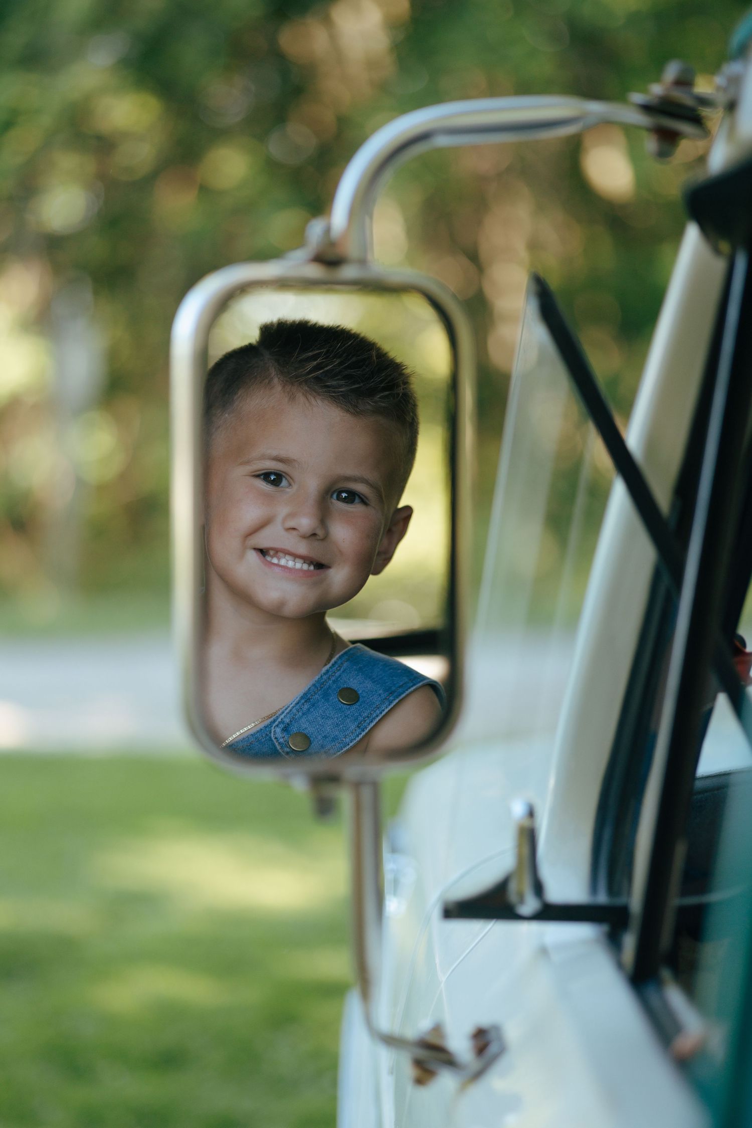Smiling boy in denim overalls reflected in vintage truck side mirror outdoors.