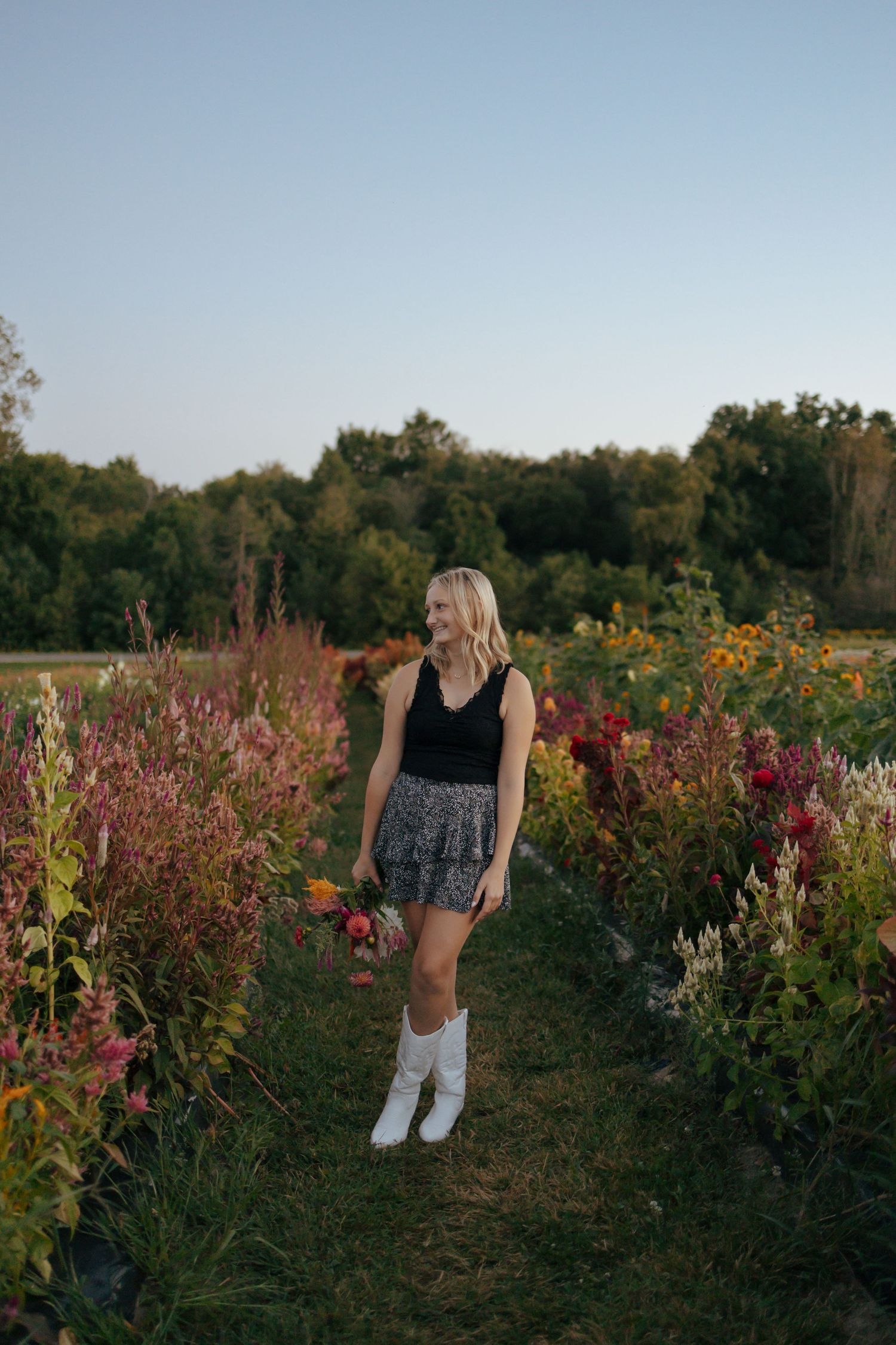 Blonde girl in black tank, floral skirt and white boots holding flowers in colorful garden at sunset.