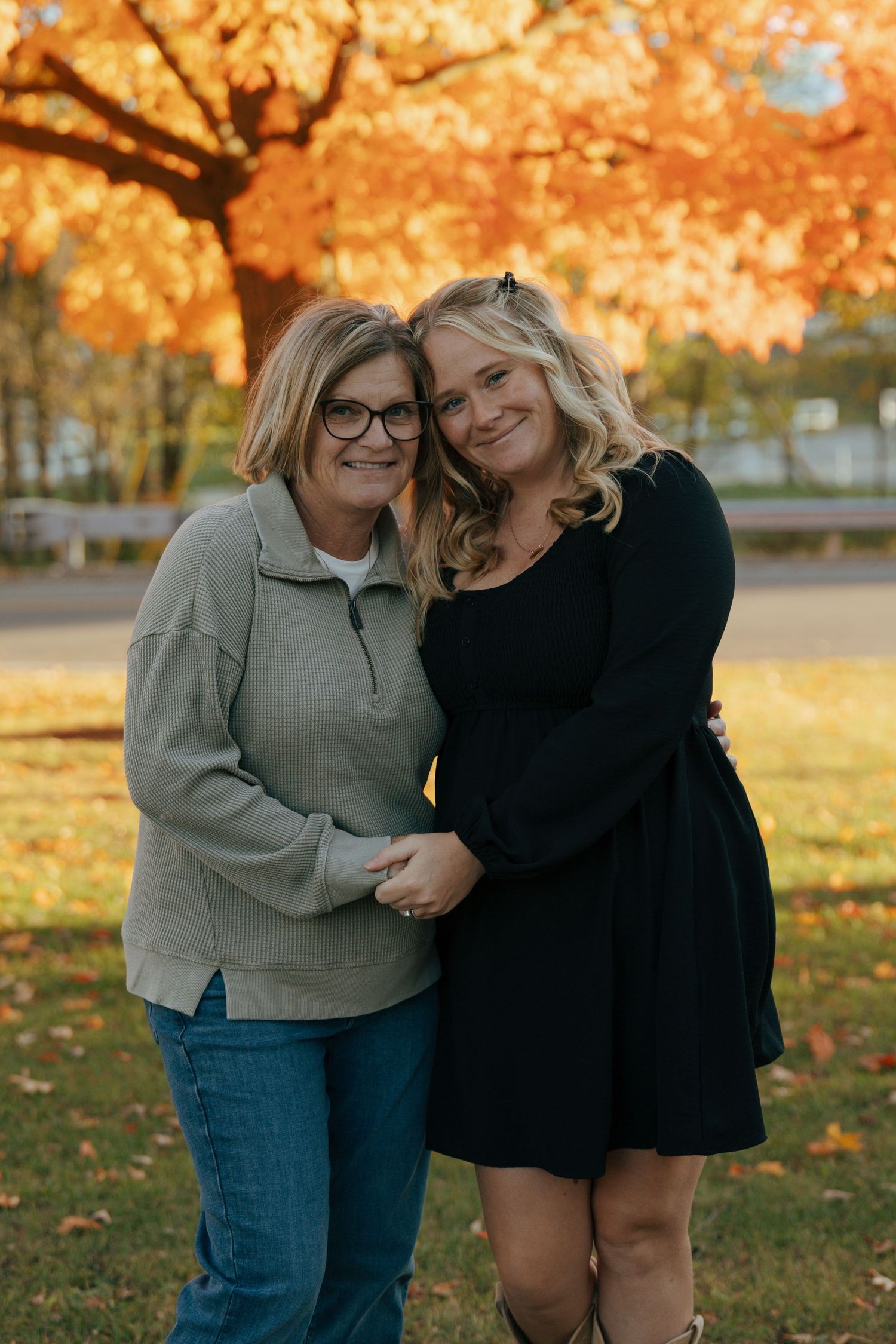 Mother and daughter smiling together in front of golden fall trees.