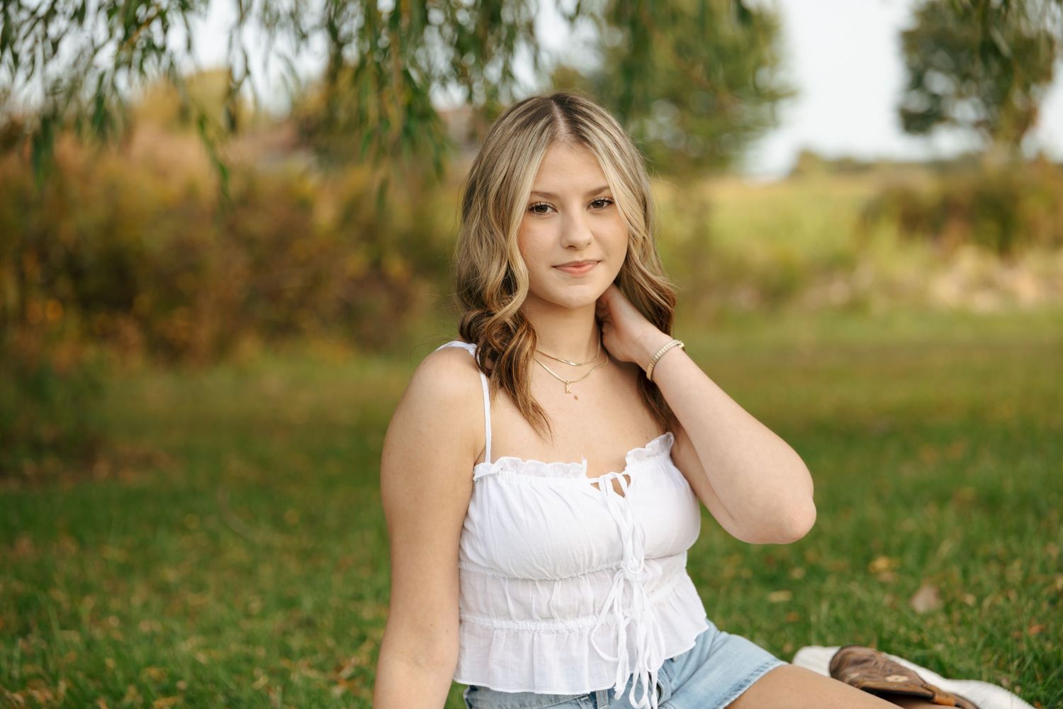 Blonde teenage girl in white crop top and denim shorts sitting on grass near weeping willow tree in golden hour light.