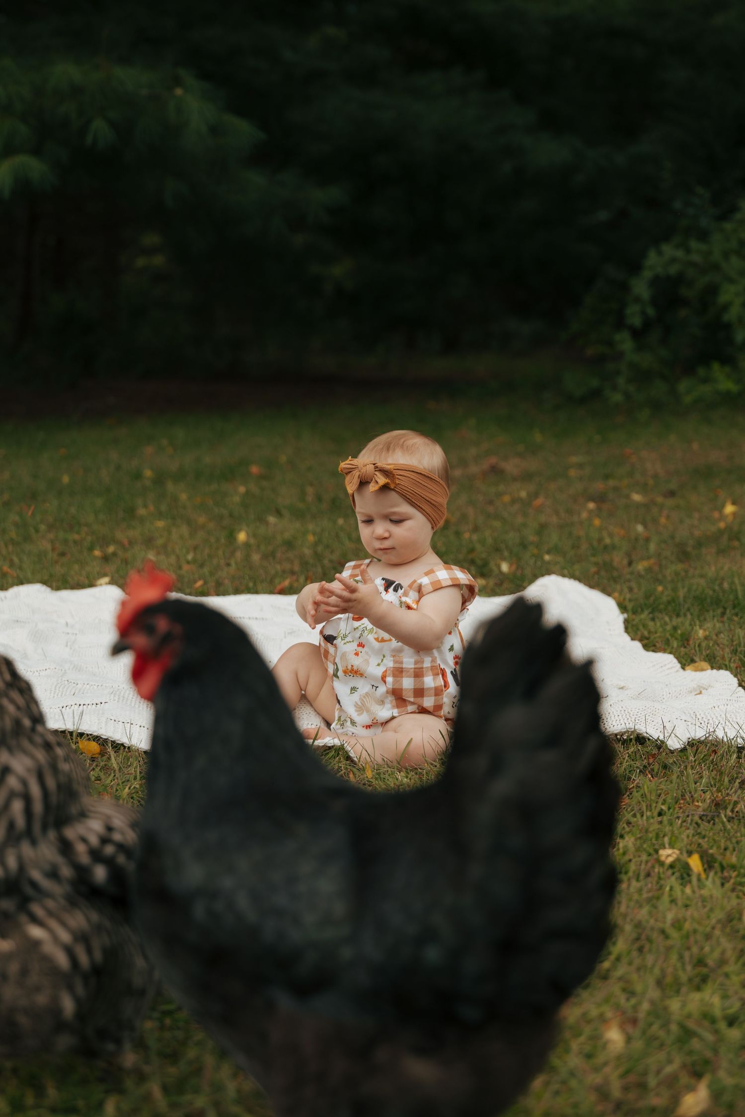 Adorable red-haired baby in plaid romper sitting on white blanket in grass surrounded by black chickens.