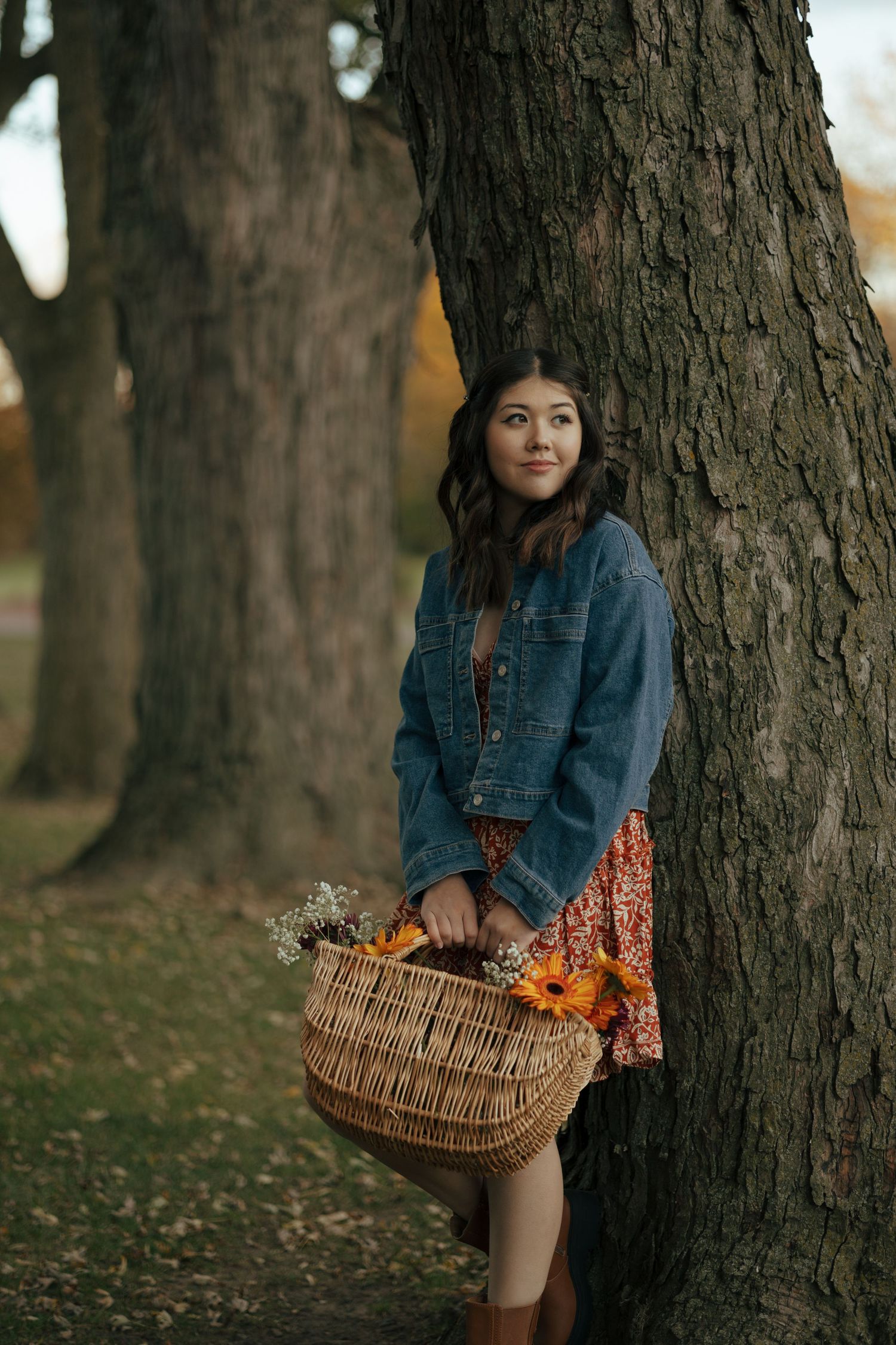 Young woman leans against large tree trunk holding a wicker basket of sunflowers in autumn park.