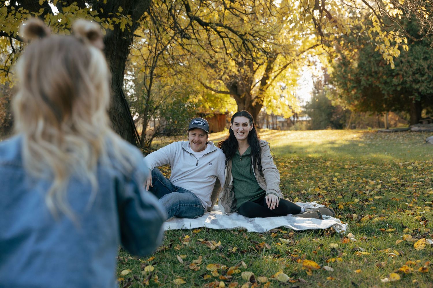 Young couple sitting on blanket under golden autumn tree smiling as their child approaches them.