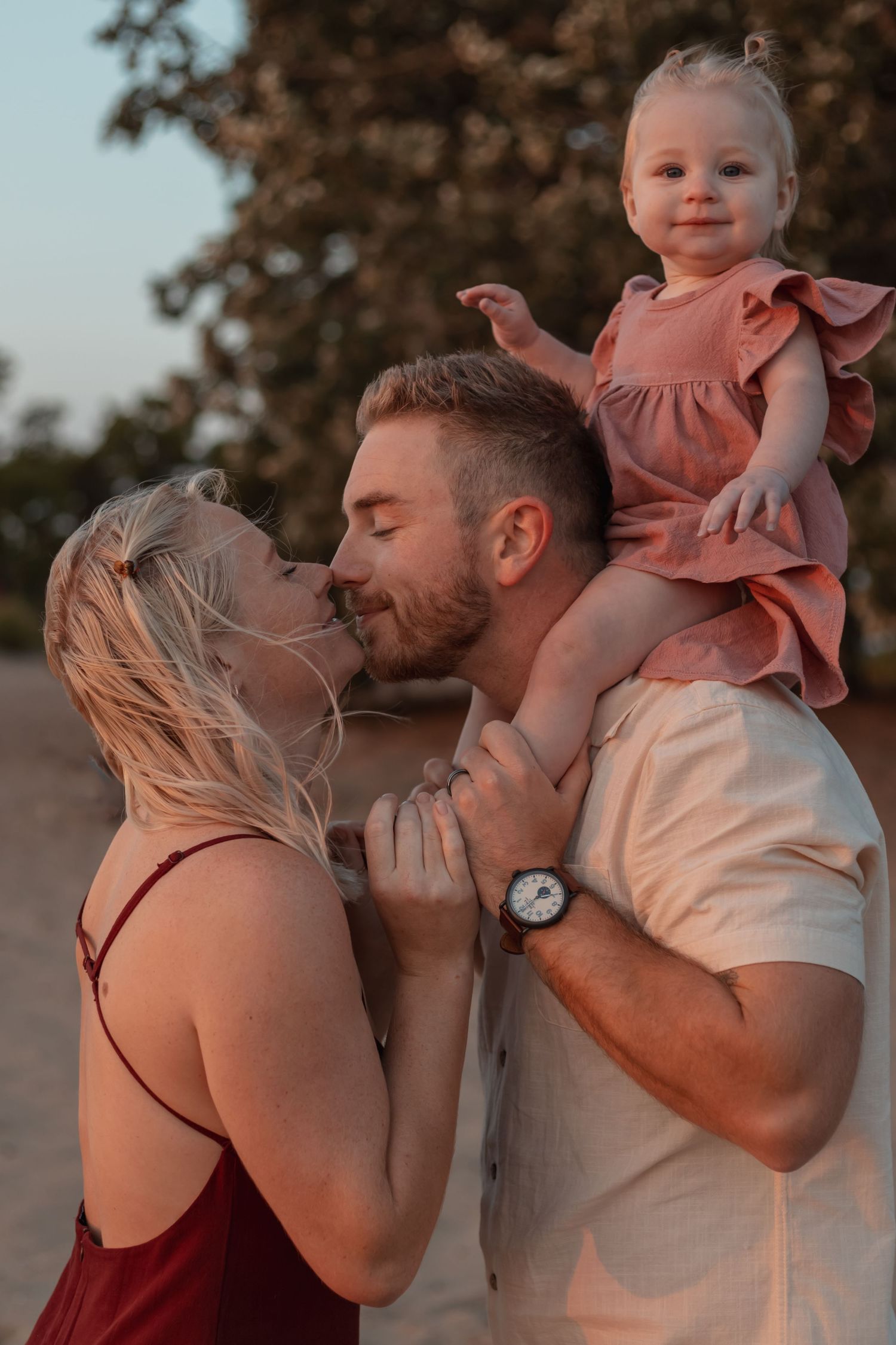 Young couple kissing while toddler girl in pink dress sits on mans shoulders outdoors.