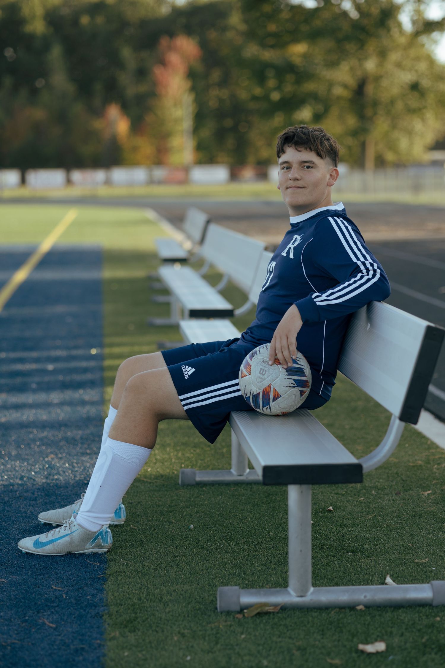 Smiling teen soccer player sits on bench holding ball in navy Adidas uniform at fall field.