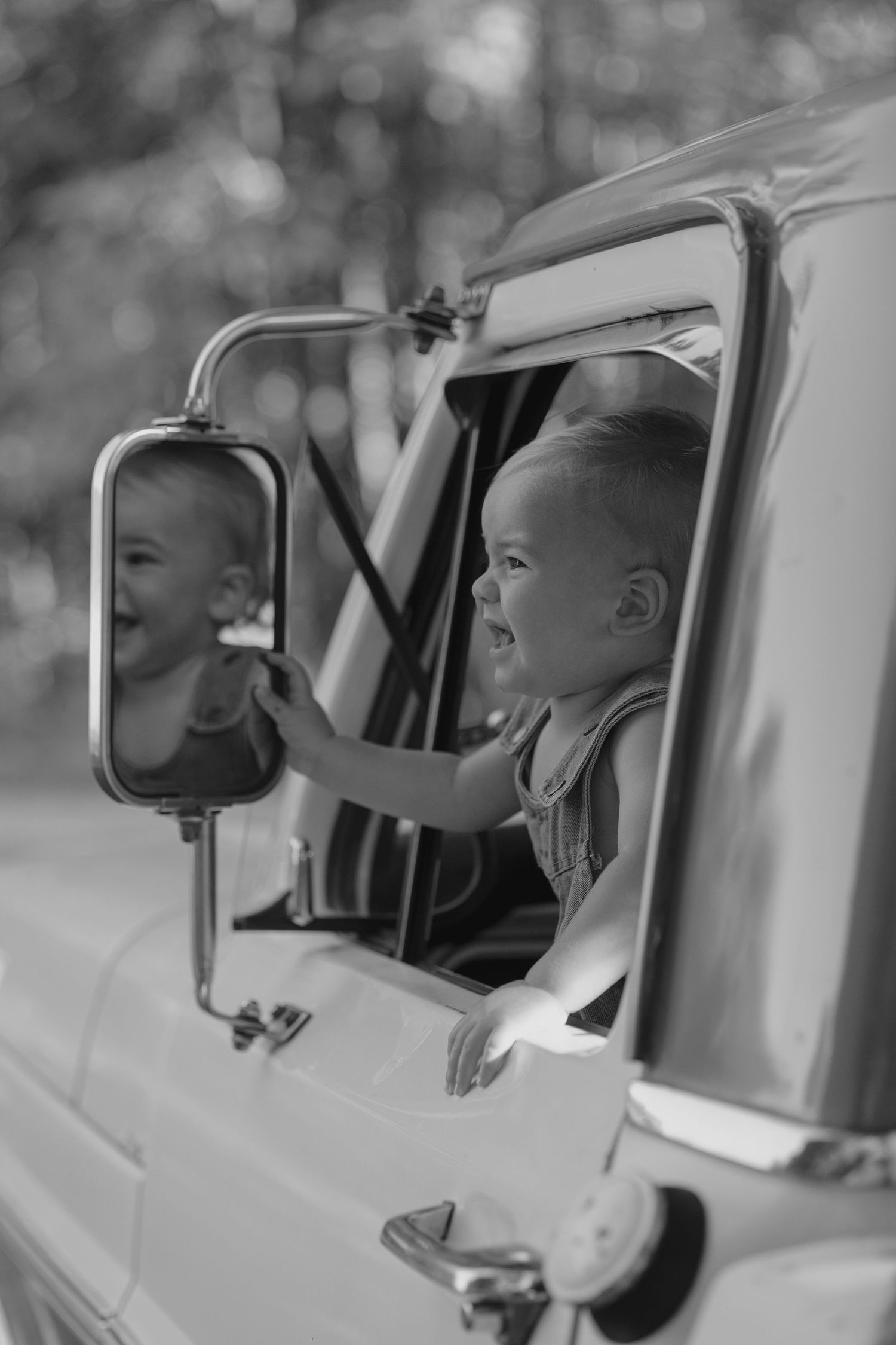 Laughing baby in denim overalls touching side mirror of vintage truck, black and white.